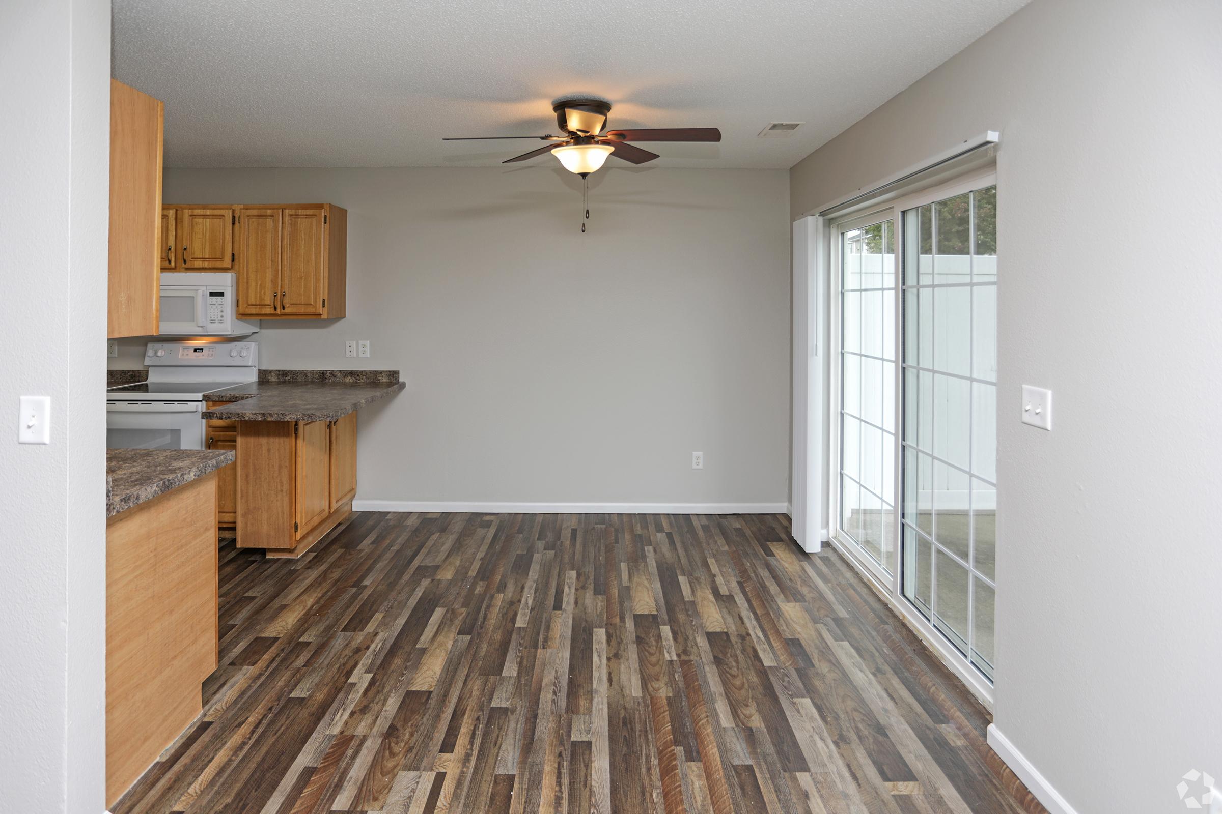A spacious, modern living area featuring hardwood-style flooring, a ceiling fan, and a large window with sliding glass doors leading to an outdoor space. The kitchen is equipped with wooden cabinets and white appliances, creating a warm and inviting atmosphere.