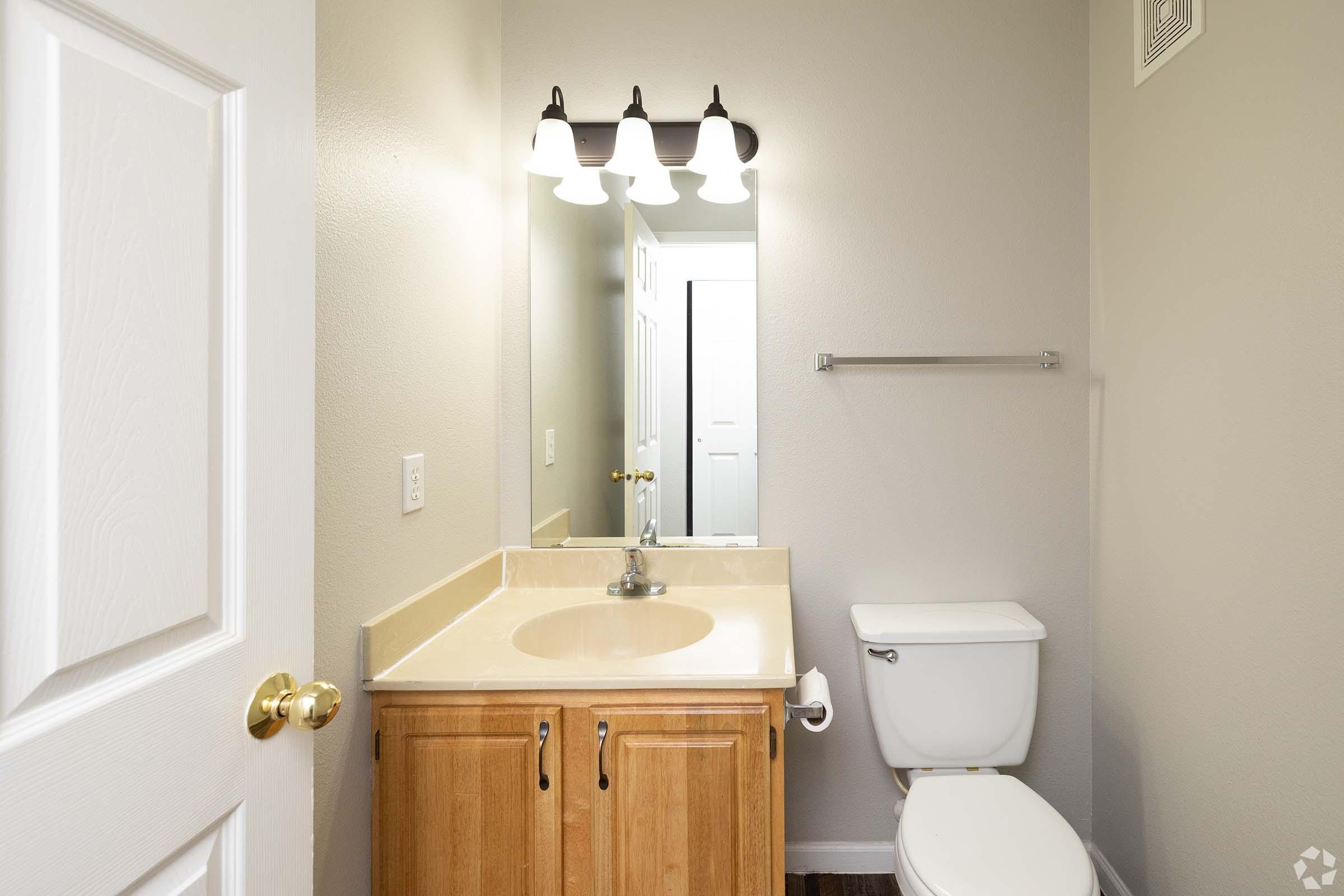 A well-lit bathroom featuring a beige vanity with a single sink, a mirror above it, and a light fixture with three bulbs. The toilet is positioned next to the vanity. The walls are painted a light color, and there is a towel bar mounted on the wall opposite the mirror.