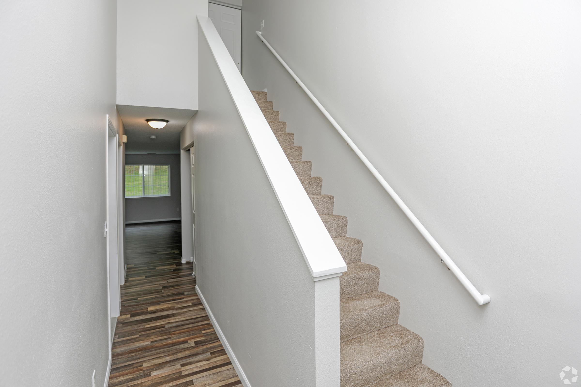 A clean and modern hallway with light gray walls and a staircase leading to the upper level. The floor features a mix of wood and carpet, and there is a door at the end of the hallway. A window allows natural light to enter, illuminating the space.