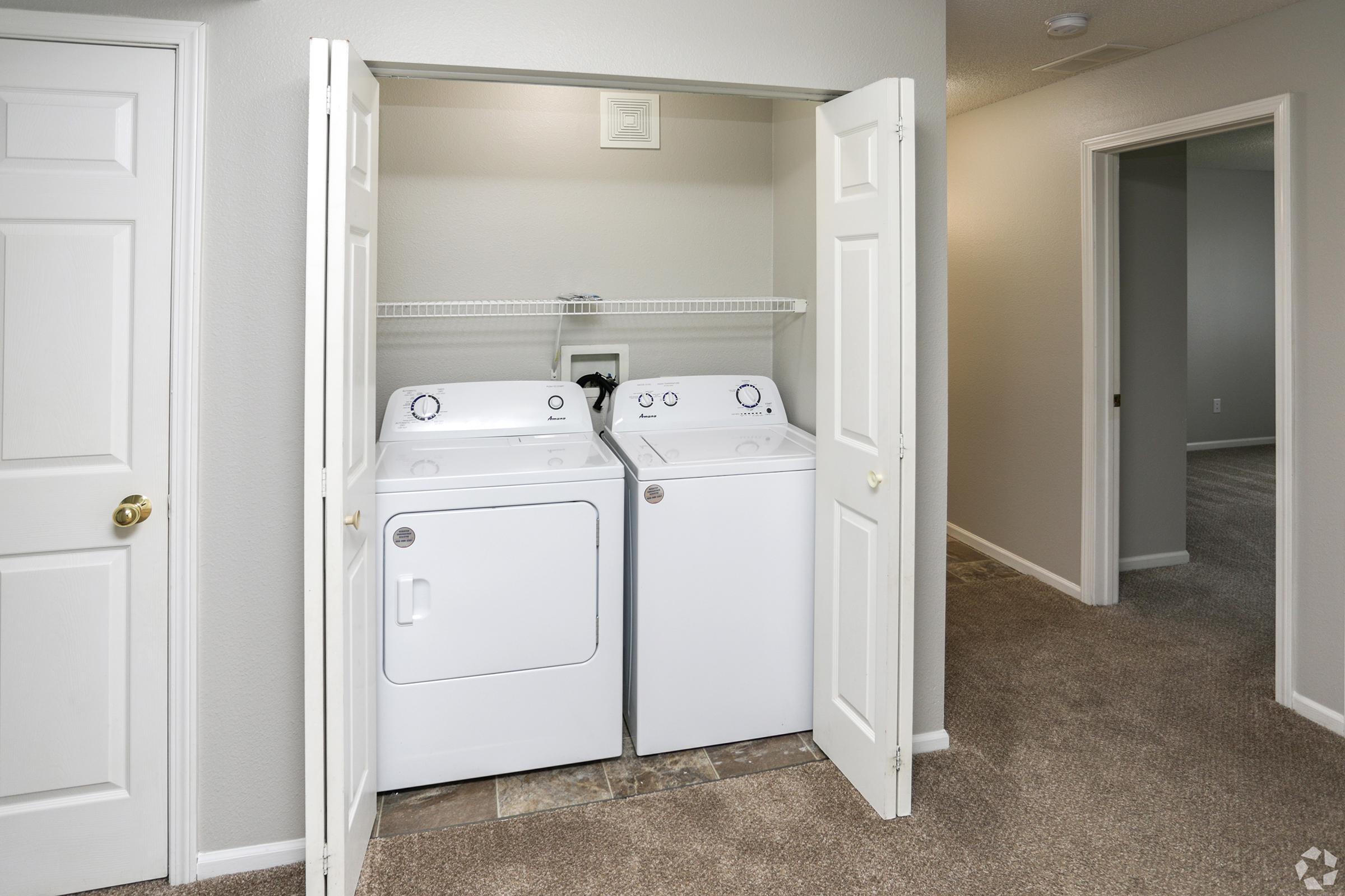 A laundry area featuring a white washer and dryer side by side, housed in a small closet with bi-fold doors opened. The surrounding area shows neutral-colored walls and carpet. There is a doorway visible on the right leading to another room, creating a spacious, organized look.