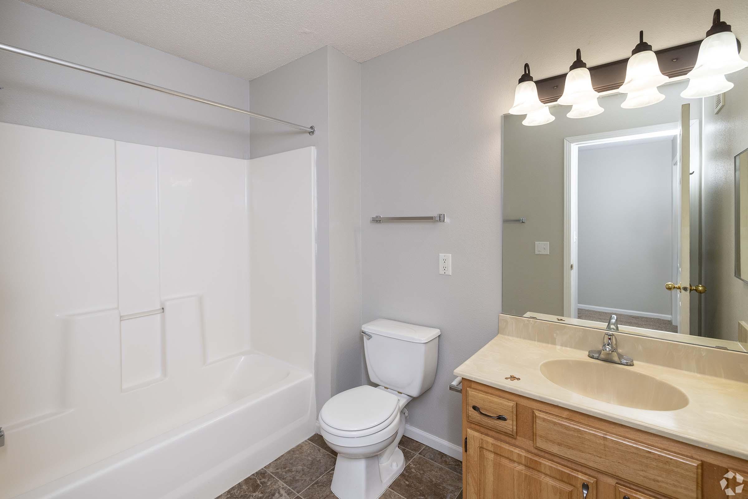 A clean and simple bathroom featuring a white bathtub and shower combination, a toilet, and a beige countertop with a sink. The walls are painted a light gray, and a mirror is mounted above the sink. Four light fixtures are attached to the wall above the mirror, providing illumination.
