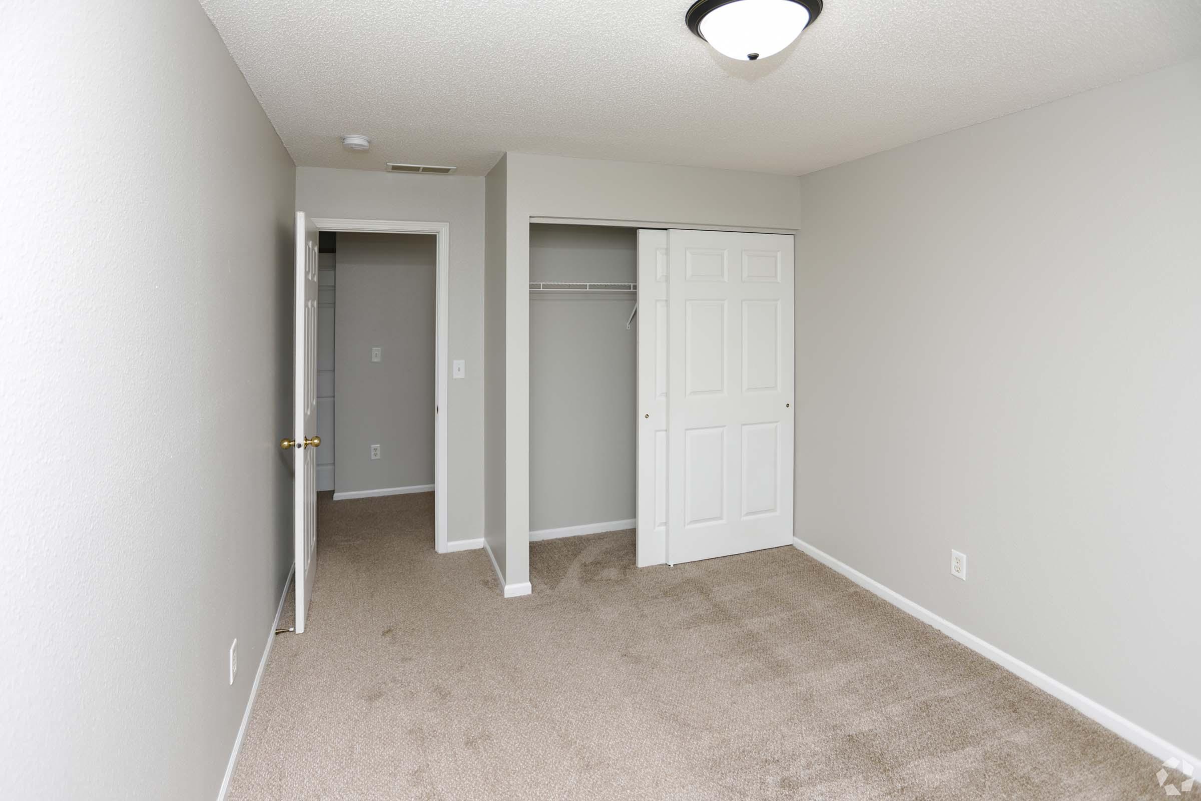Empty room with light gray walls and carpet. One wall features a closed double door closet. In the background, there's an open door leading to another small room. The ceiling has a round light fixture. Overall, the space is clean and well-lit, creating a minimalist aesthetic.