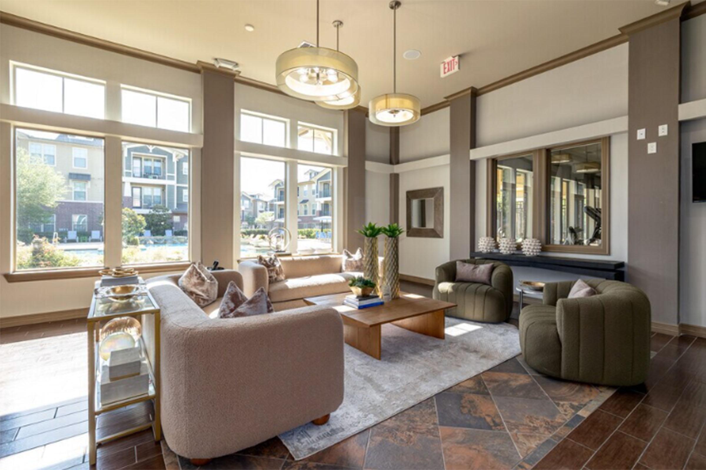 A modern lounge area featuring a large seating arrangement with plush sofas, a coffee table, and decorative pillows. Large windows allow natural light to fill the space, offering views of an outdoor area. Stylish lighting fixtures hang from the ceiling, and potted plants add a touch of greenery.