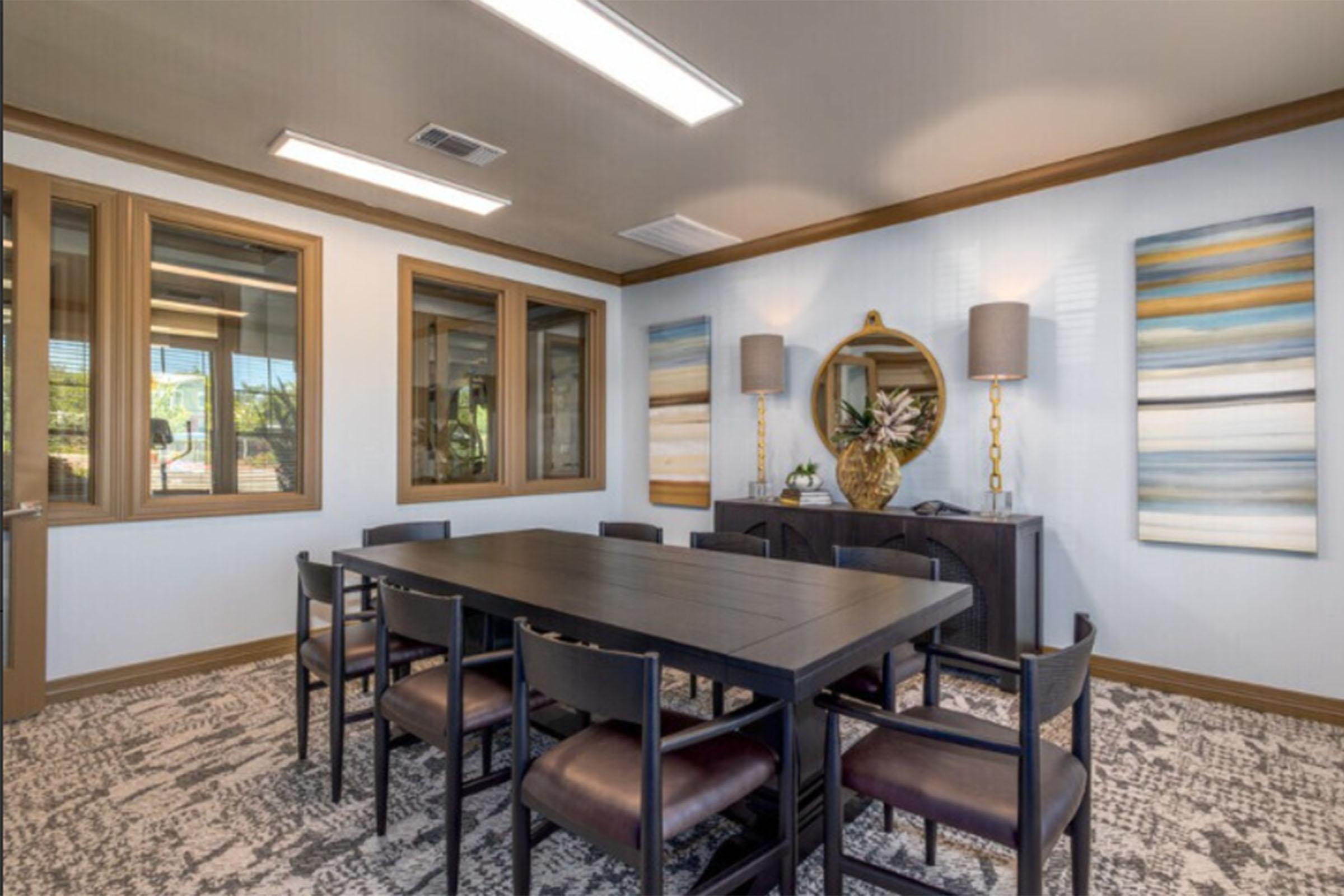 A modern meeting room featuring a large black dining table surrounded by eight chairs. The walls are adorned with two abstract paintings and a round mirror. There are two stylish lamps on either side of a decorative vase with plants. Large windows allow natural light to brighten the space.