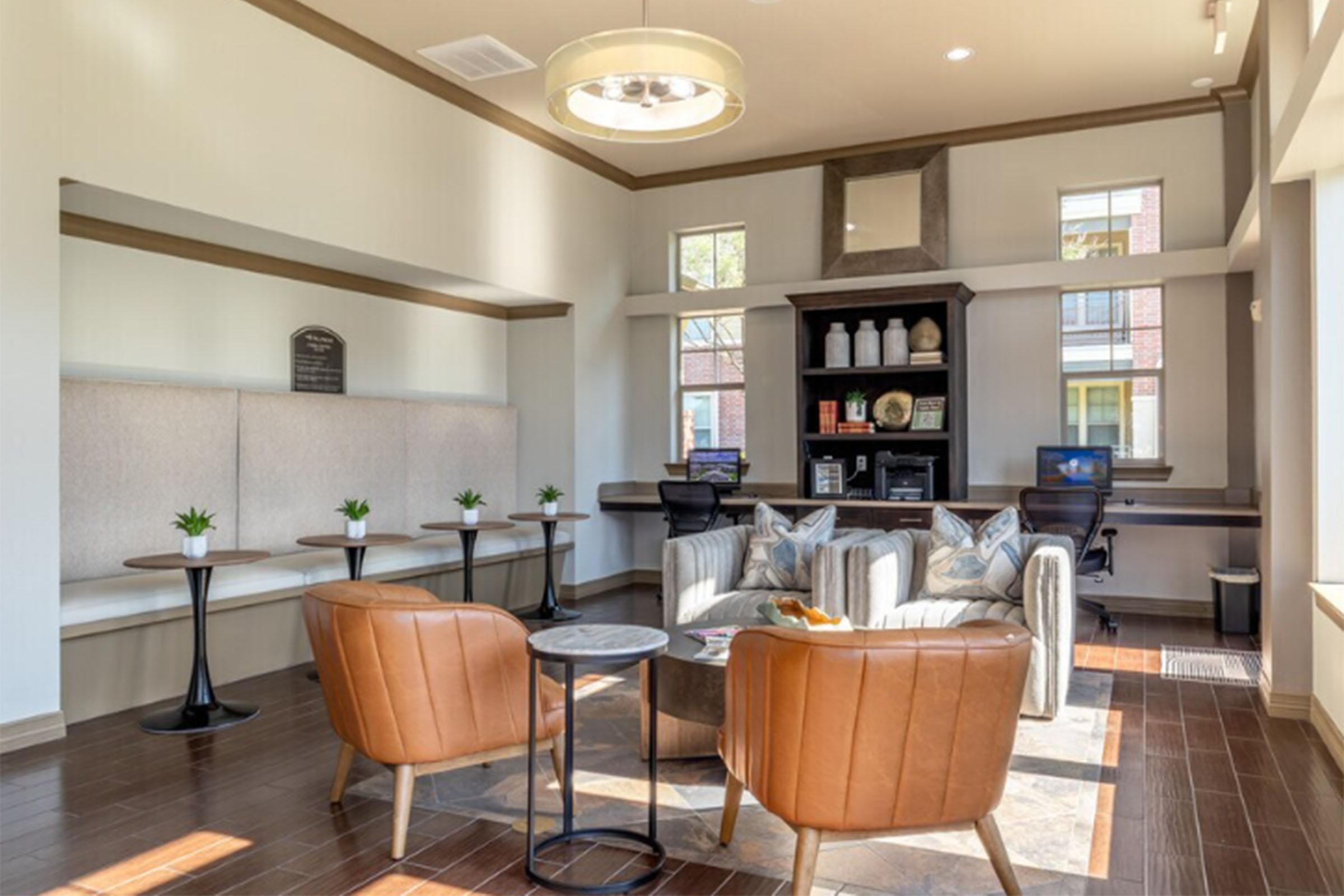 A modern lounge area featuring two brown leather chairs, a small round table, and a cozy design aesthetic. The room has light-colored walls, a decorative bookshelf, large windows allowing natural light, and plants for a welcoming atmosphere, ideal for relaxation or socializing.