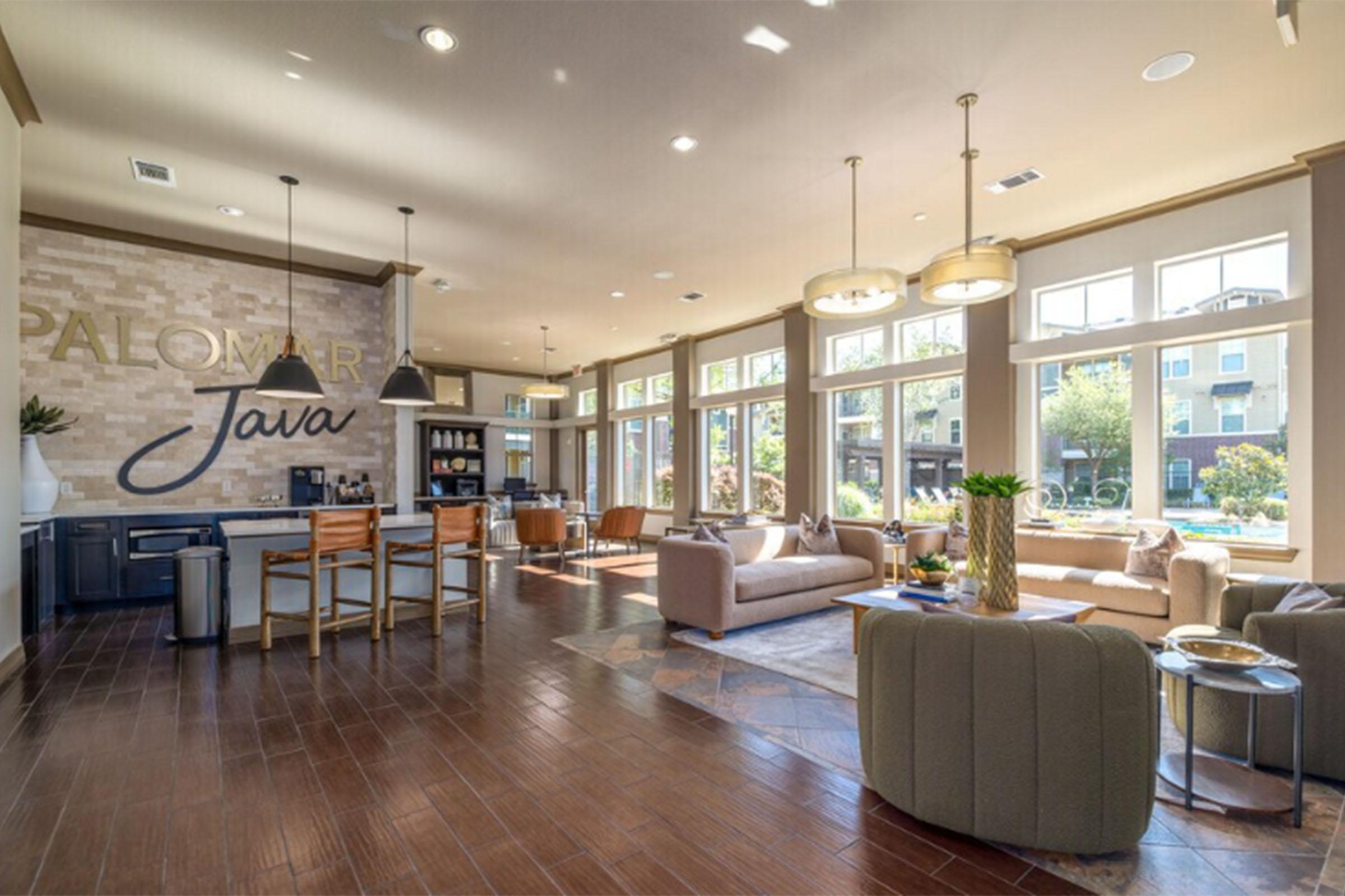 A spacious, modern lounge area with large windows allowing natural light. Features include a coffee station, comfortable seating arrangements with sofas and chairs, decorative plants, and stylish lighting fixtures. The walls are adorned with the words "PALOMAR Java" and a cozy, inviting atmosphere is created.