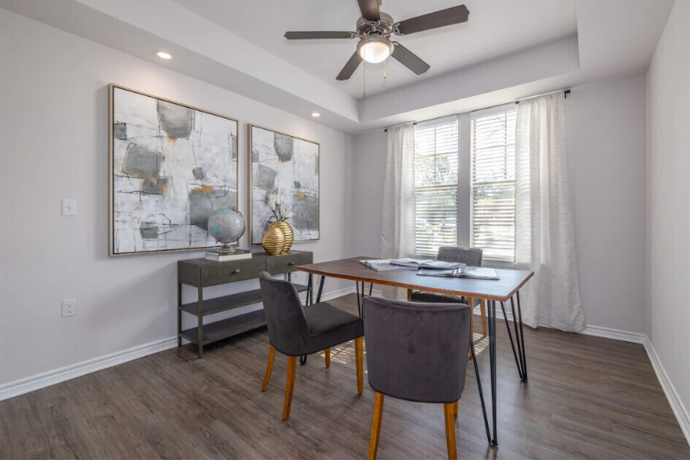 A modern home office featuring a wooden desk with a table lamp, two gray chairs, and decorative items. The room has large windows with white curtains, a ceiling fan, and two abstract paintings on the wall. The floor is made of light wood, creating a clean and inviting workspace atmosphere.