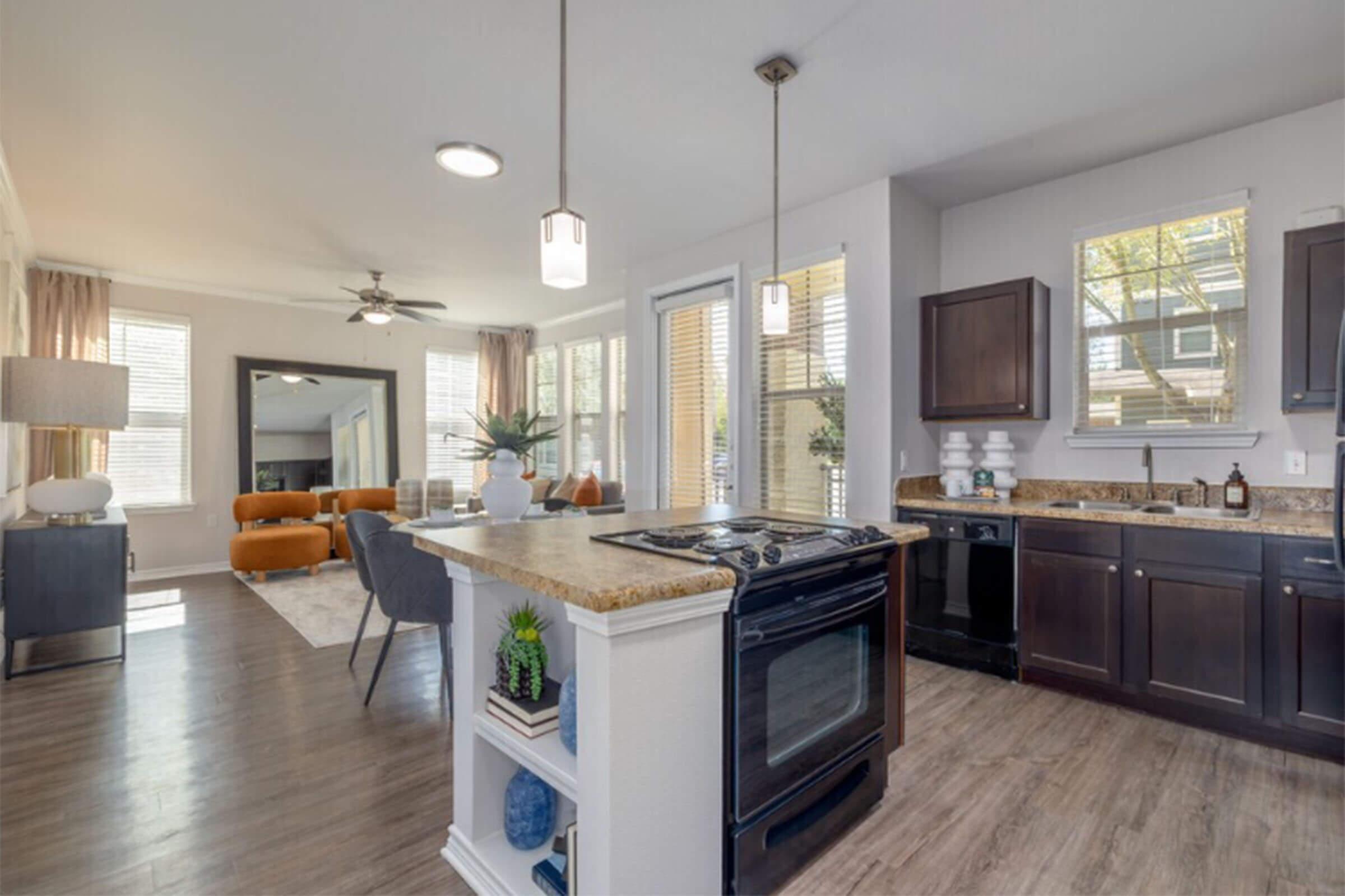 A modern open-concept kitchen and living area featuring granite countertops, dark wood cabinetry, and stainless steel appliances. The living space includes a comfortable sofa, a large window with natural light, and stylish decor, creating a cozy and inviting atmosphere.