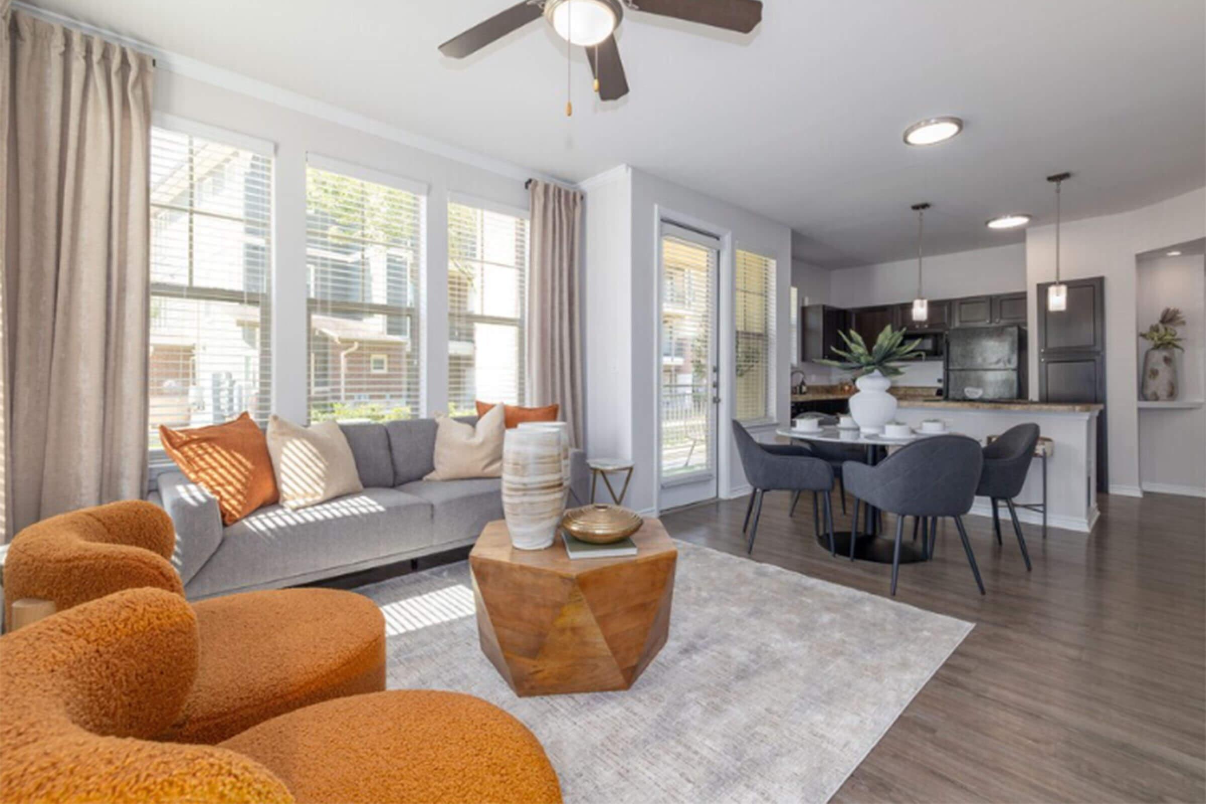 A stylish living room featuring a gray sofa adorned with colorful pillows, two plush orange chairs, and a wooden coffee table. Natural light streams in through large windows, illuminating a dining area with a round table and black chairs. Modern decor elements complement the inviting atmosphere.