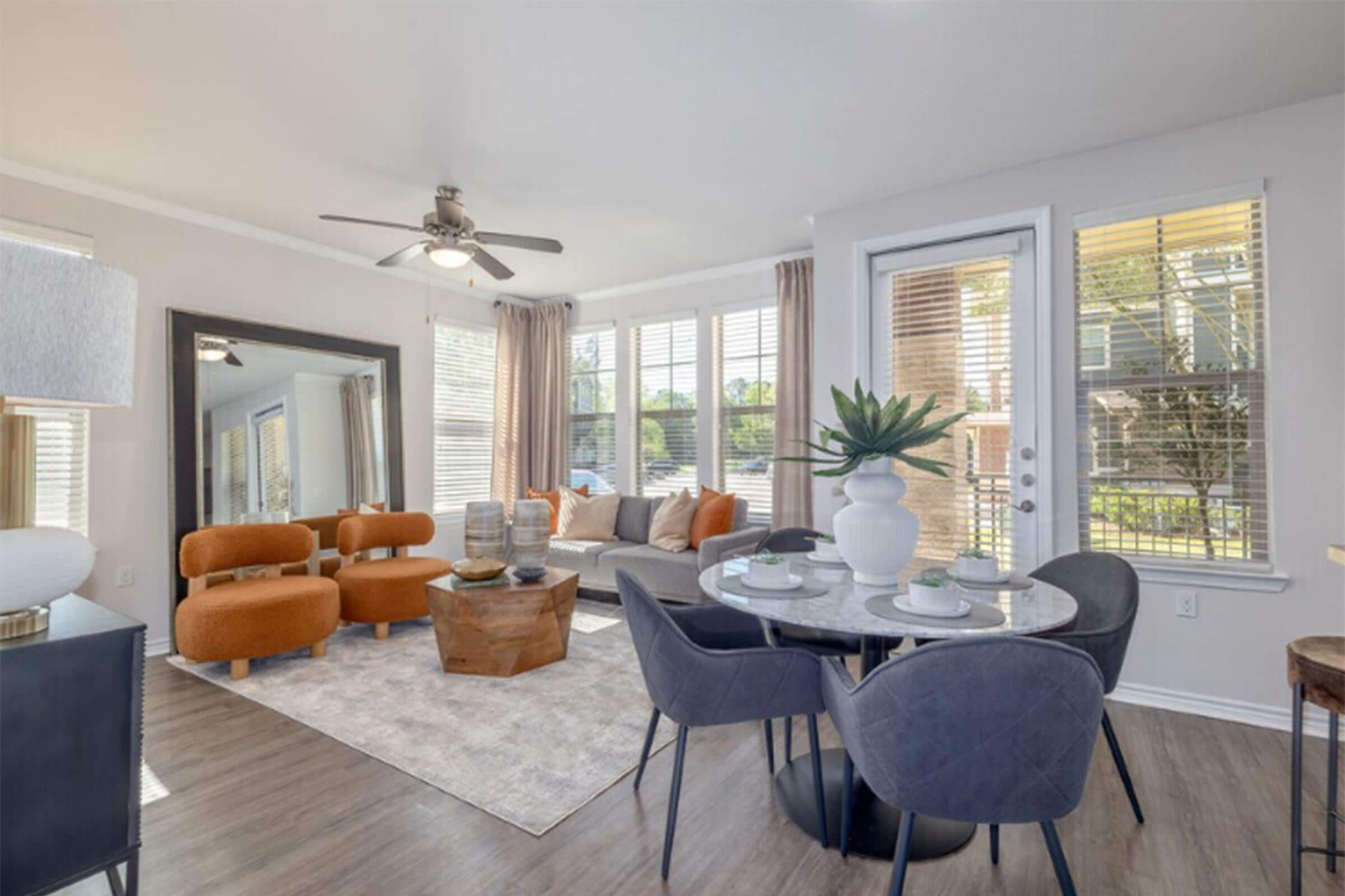 A modern living room featuring a cozy seating area with orange chairs, a round dining table with four chairs, and a large mirror. Sunlight pours in through multiple windows, highlighting a decorative plant and a stylish rug that adds warmth to the space.