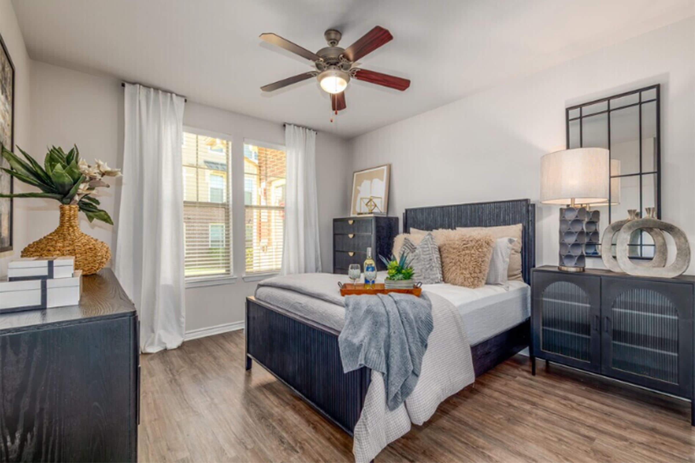 A modern bedroom featuring a bed with decorative pillows and a gray blanket. There's a nightstand with a lamp, decorative items, and a window with white curtains. A dresser and a black cabinet are in the room, along with a ceiling fan and a vase with flowers. The wood flooring adds warmth to the space.