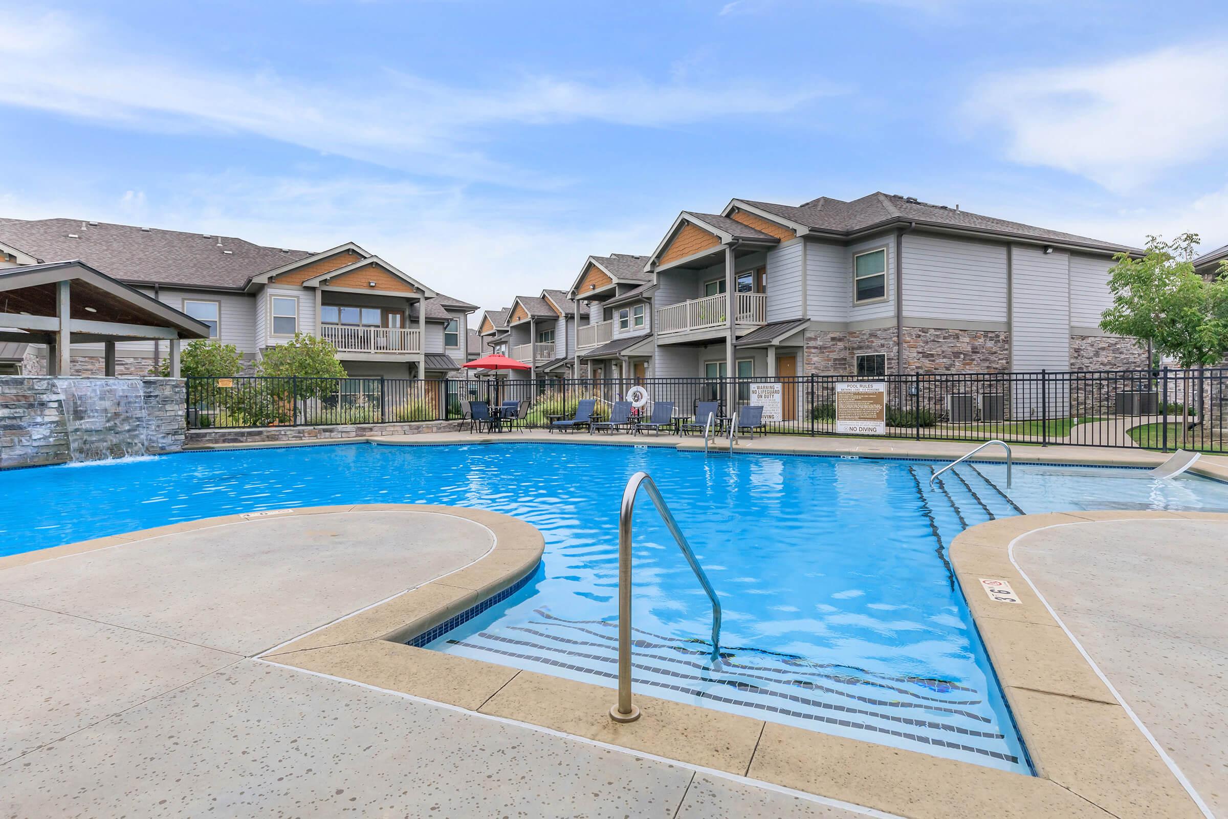 A large, inviting swimming pool at an apartment complex, surrounded by lounge chairs and umbrellas. In the background, there are modern buildings with balconies and landscaped greenery under a clear blue sky. The area provides a relaxing atmosphere for residents and guests.