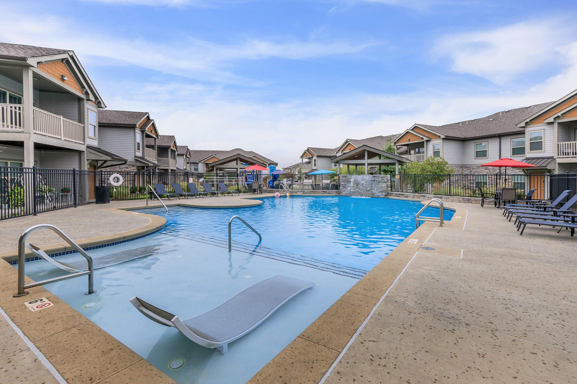 A spacious outdoor swimming pool surrounded by lounge chairs and shaded umbrellas, located within an apartment complex. The area features colorful slides, landscaped greenery, and multiple two-story buildings in the background under a clear blue sky.