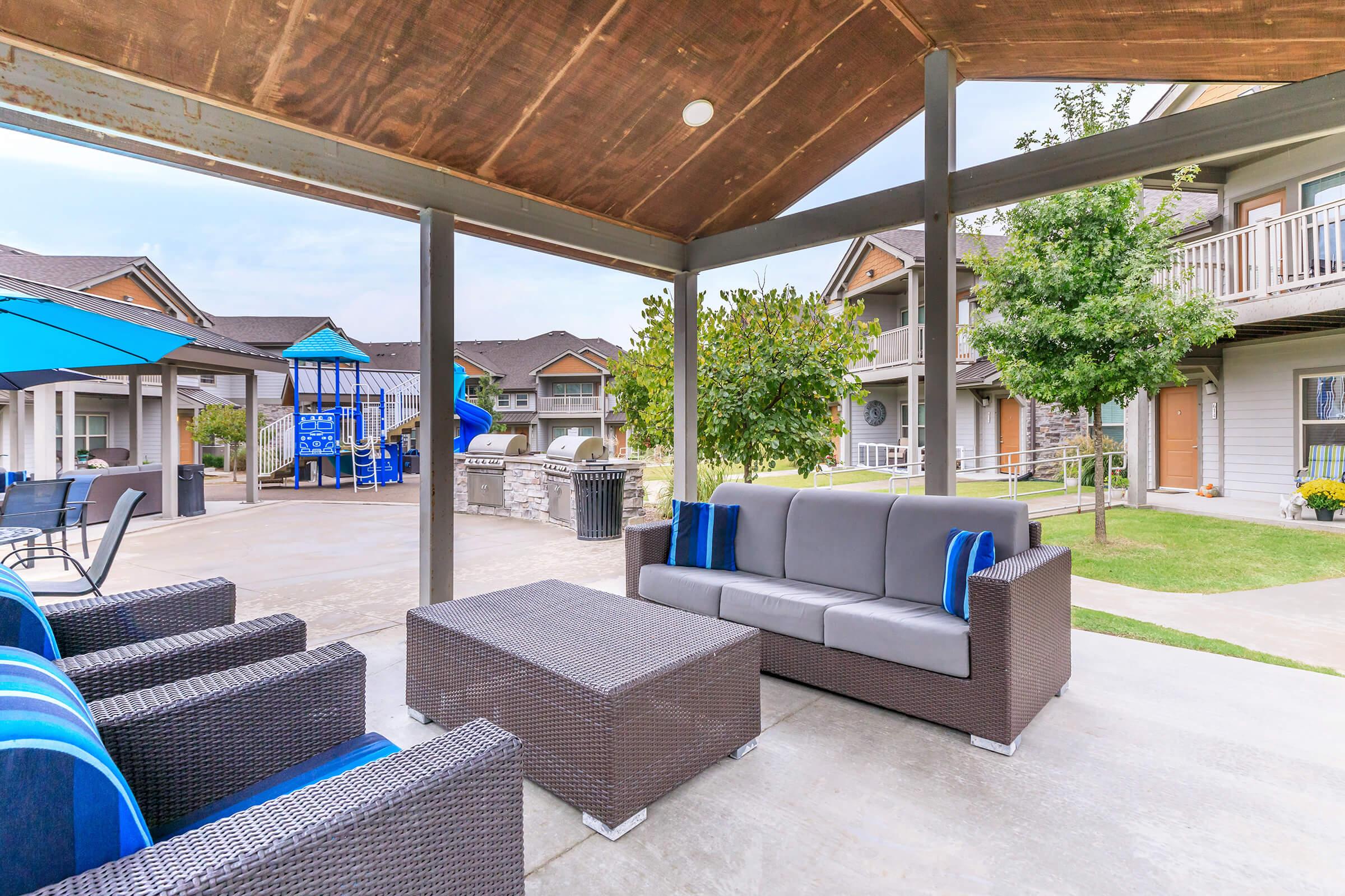 A shaded outdoor seating area featuring a gray wicker sofa with blue cushions and a matching coffee table. Nearby, there are additional wicker chairs and blue umbrellas. In the background, there are residential buildings and a playground with slides and climbing structures, surrounded by green grass and trees.