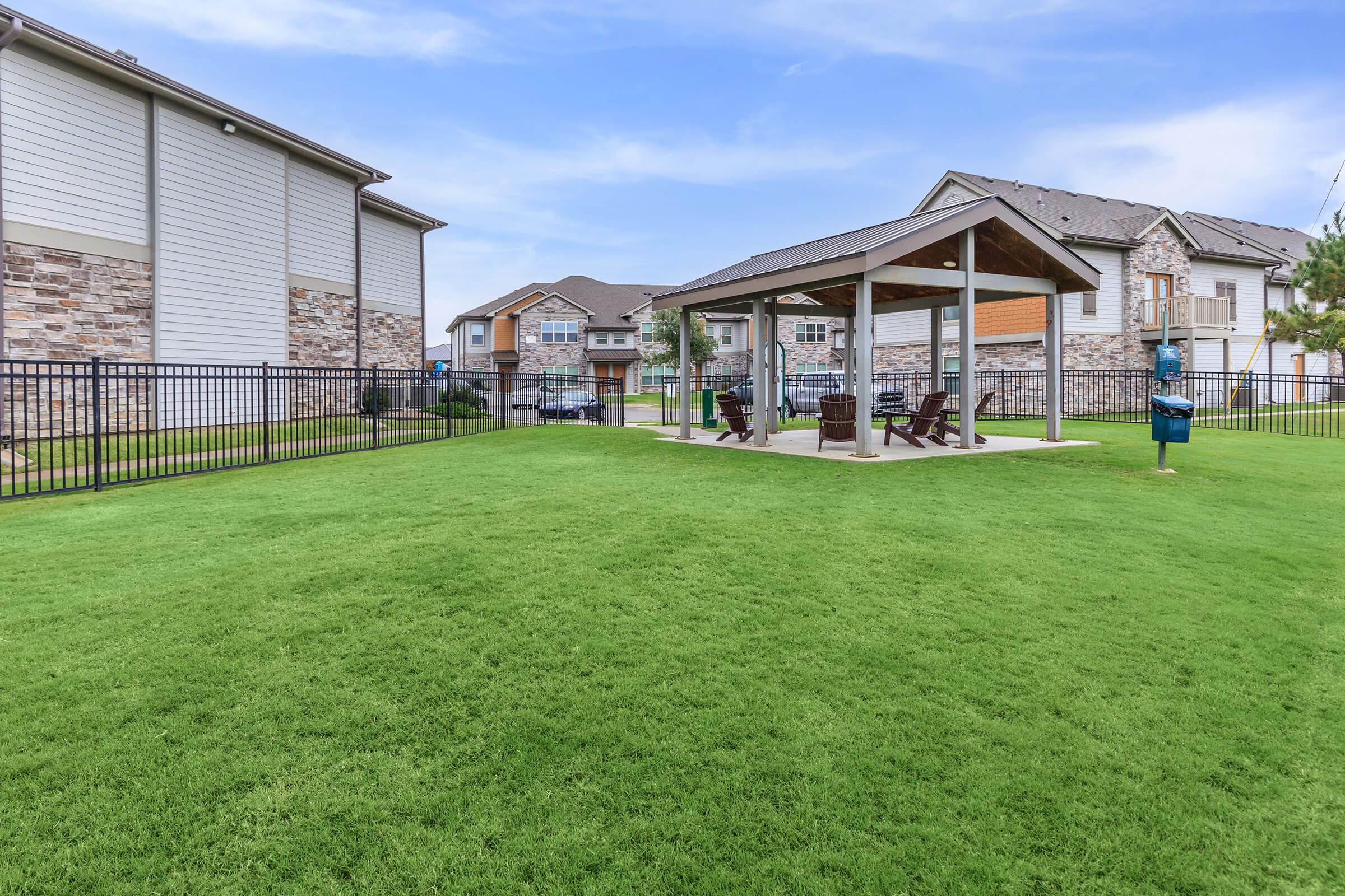 A well-maintained grass area featuring a covered gazebo with wooden seating. Surrounding the gazebo is a fenced yard, and several buildings can be seen in the background. The sky is clear, creating a serene outdoor space suitable for relaxation and gatherings.