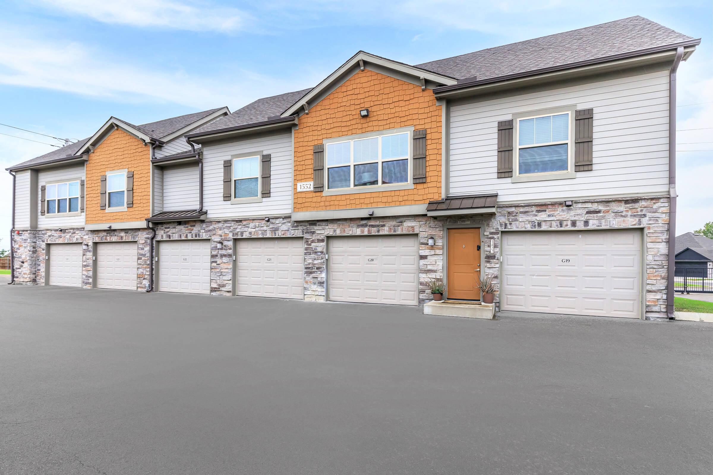 Row of modern townhouses featuring beige and orange exteriors, large windows, and stone accents. Each unit has a garage door and a small front porch, with a neatly paved driveway and blue sky in the background.