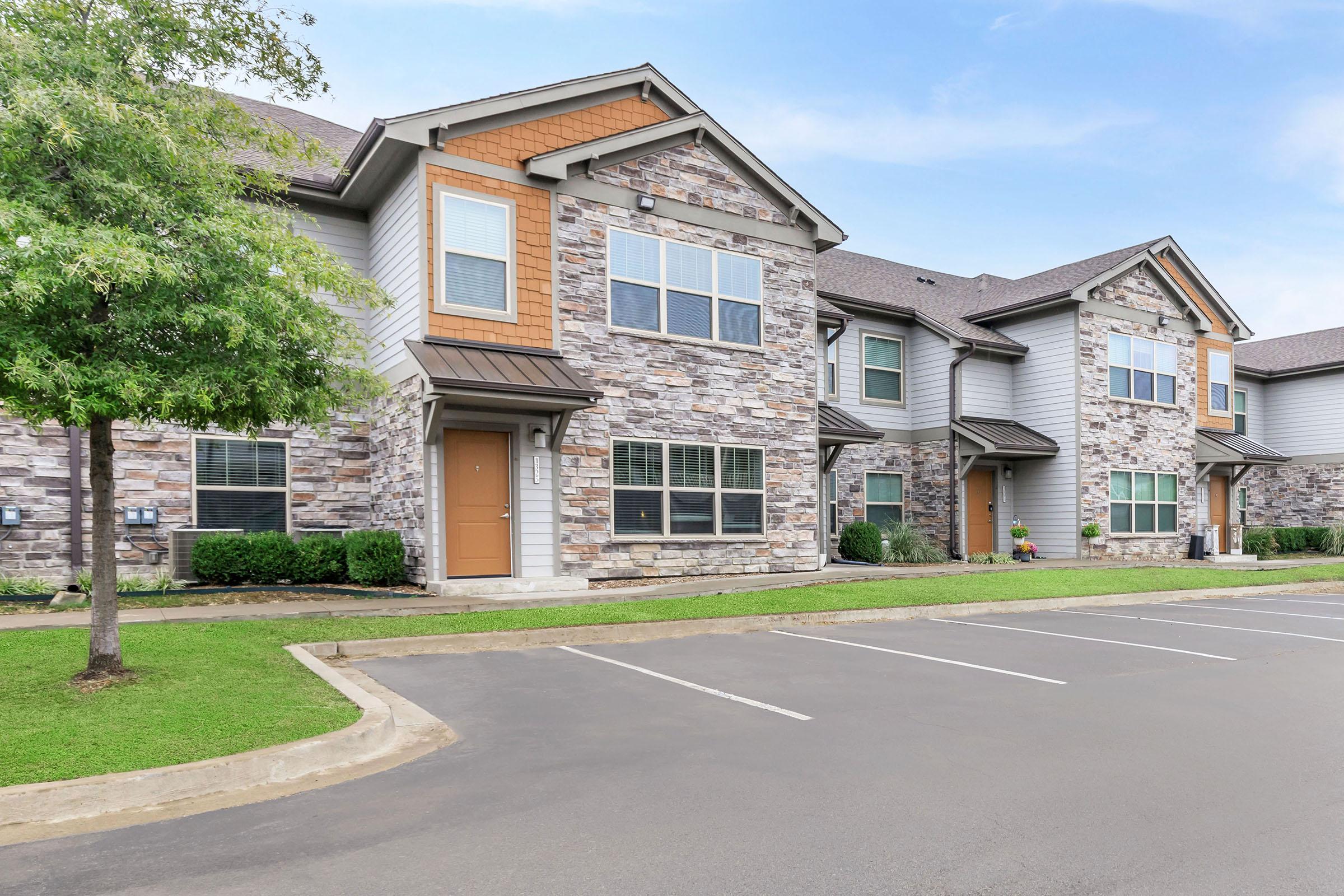 A modern apartment complex featuring two-story buildings with stone and siding exteriors. The foreground shows a well-maintained lawn and a parking area with empty spaces. The buildings have large windows and front doors, creating an inviting atmosphere in a suburban setting.