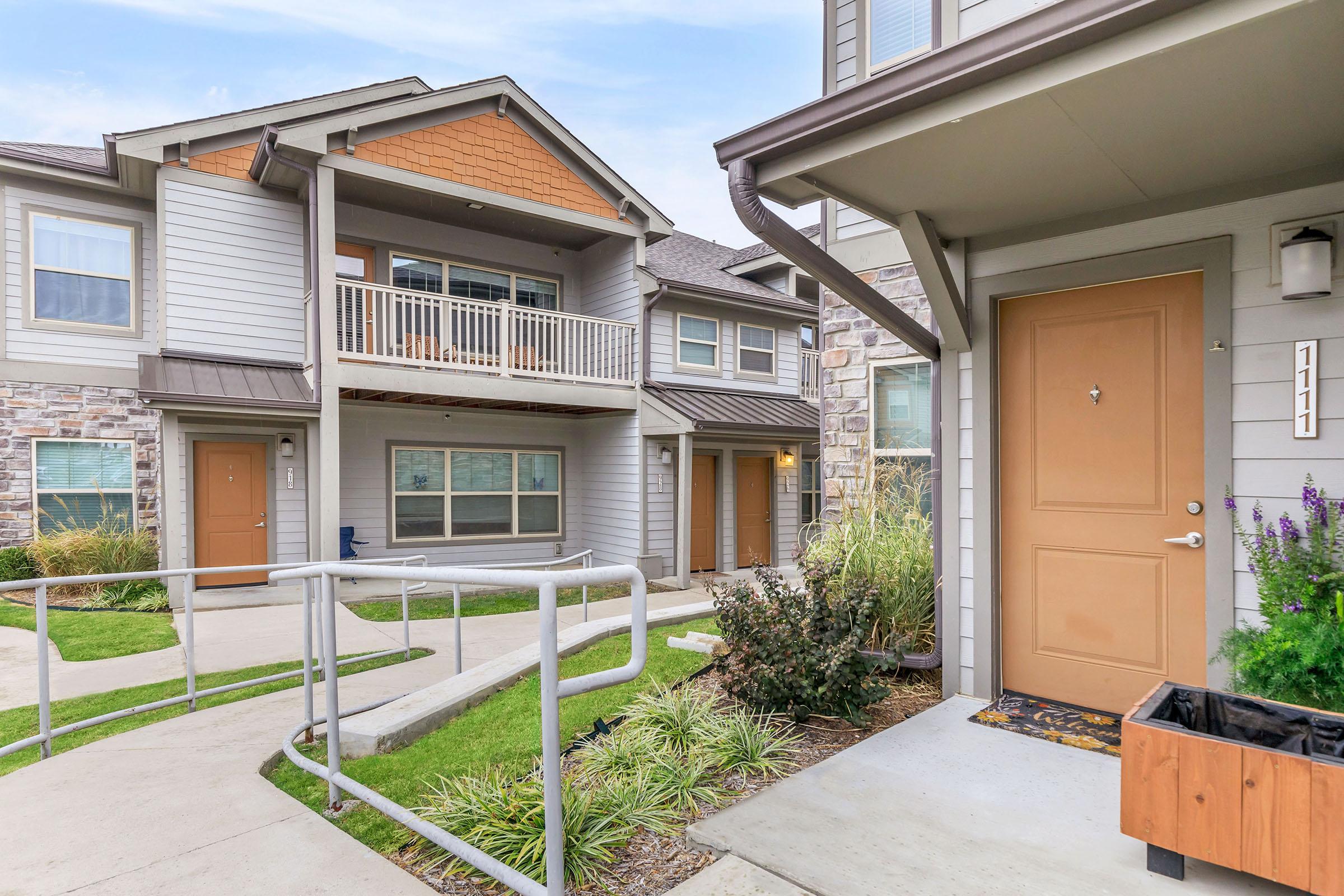 A view of a multi-story apartment complex featuring a light gray exterior, brown accents, and a welcoming entrance with a door. The landscape includes well-maintained lawns, shrubs, and a ramp for accessibility. Balconies are visible on the upper levels, enhancing the inviting aesthetic of the area.