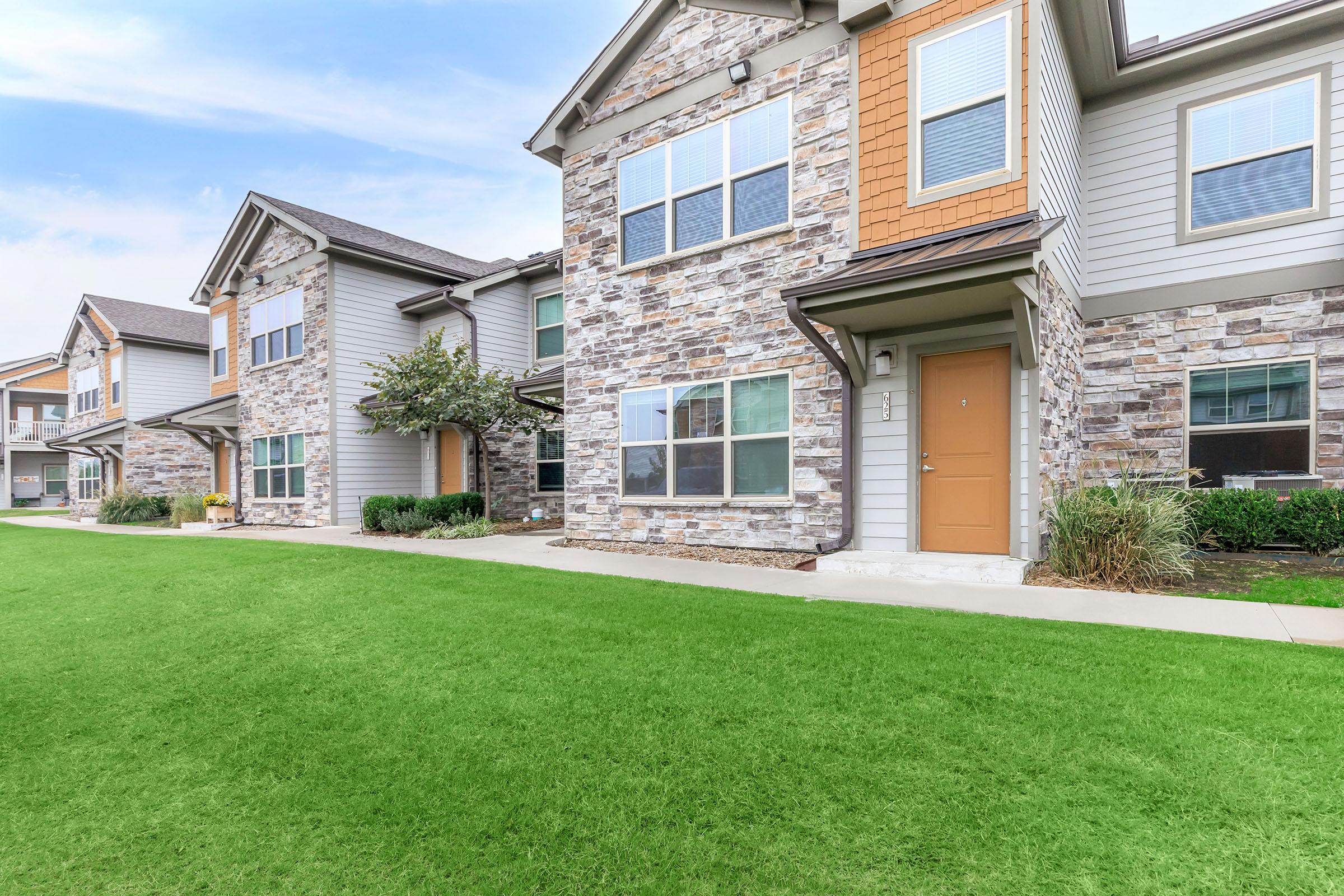 A row of modern two-story apartment buildings featuring a blend of stone and siding exteriors. Each unit has large windows and a front door, surrounded by well-manicured green lawns. Trees and shrubs enhance the landscaping, creating an inviting atmosphere in a residential community.