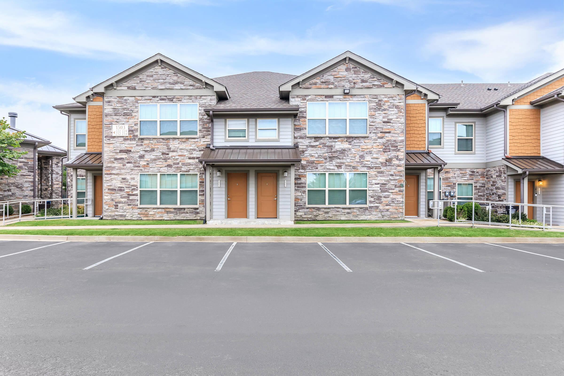 A modern two-story apartment building featuring a stone exterior with multiple windows and a gabled roof. The front entrance has double doors, and there is a neatly landscaped area with a small lawn. Adjacent parking spaces are visible in the foreground, indicating a residential community.