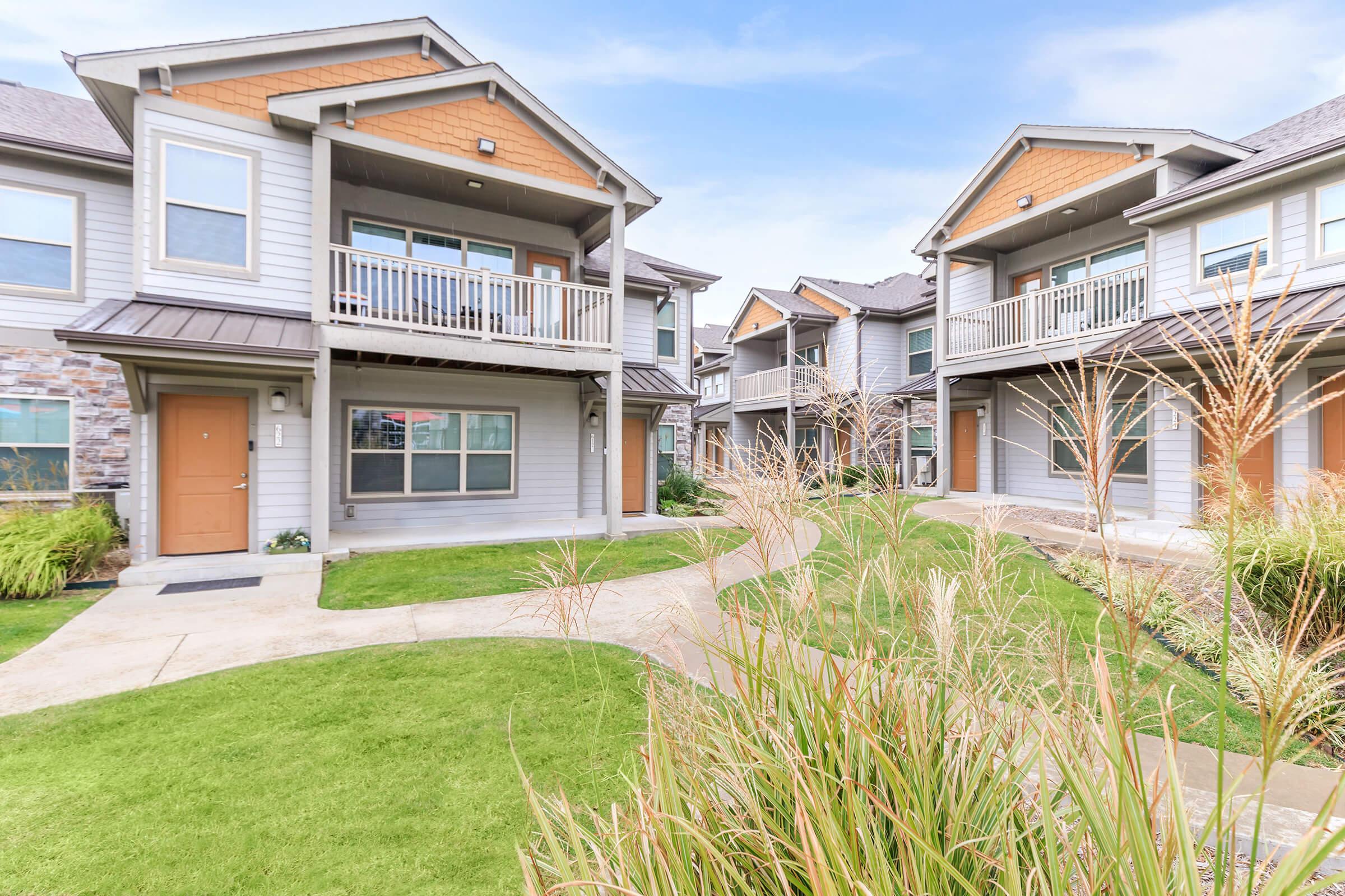 A view of a modern residential complex featuring several two-story buildings with balconies. The architecture includes a mix of stone and siding. Lush green grass and landscaped areas with ornamental grasses surround the walkway leading to the entrances of the units. The sky is clear and blue.
