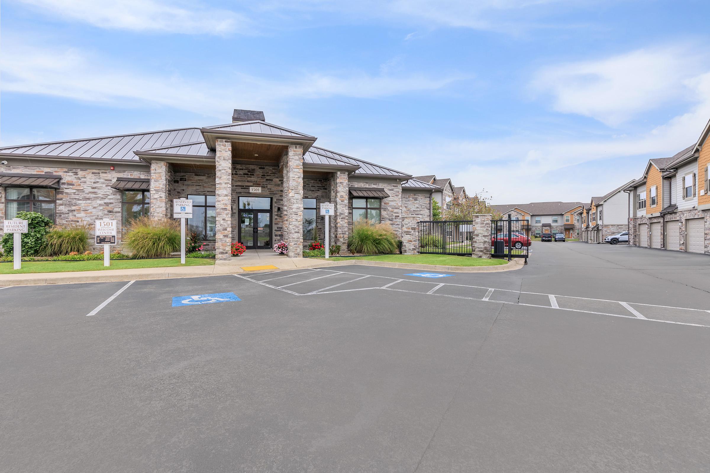 A modern stone-clad building with a sloped roof, featuring large windows and a welcoming entrance. The landscaped area includes greenery and flowers. In the foreground, there are parking spaces, including designated spots for handicapped parking. Additional residential buildings are visible in the background.