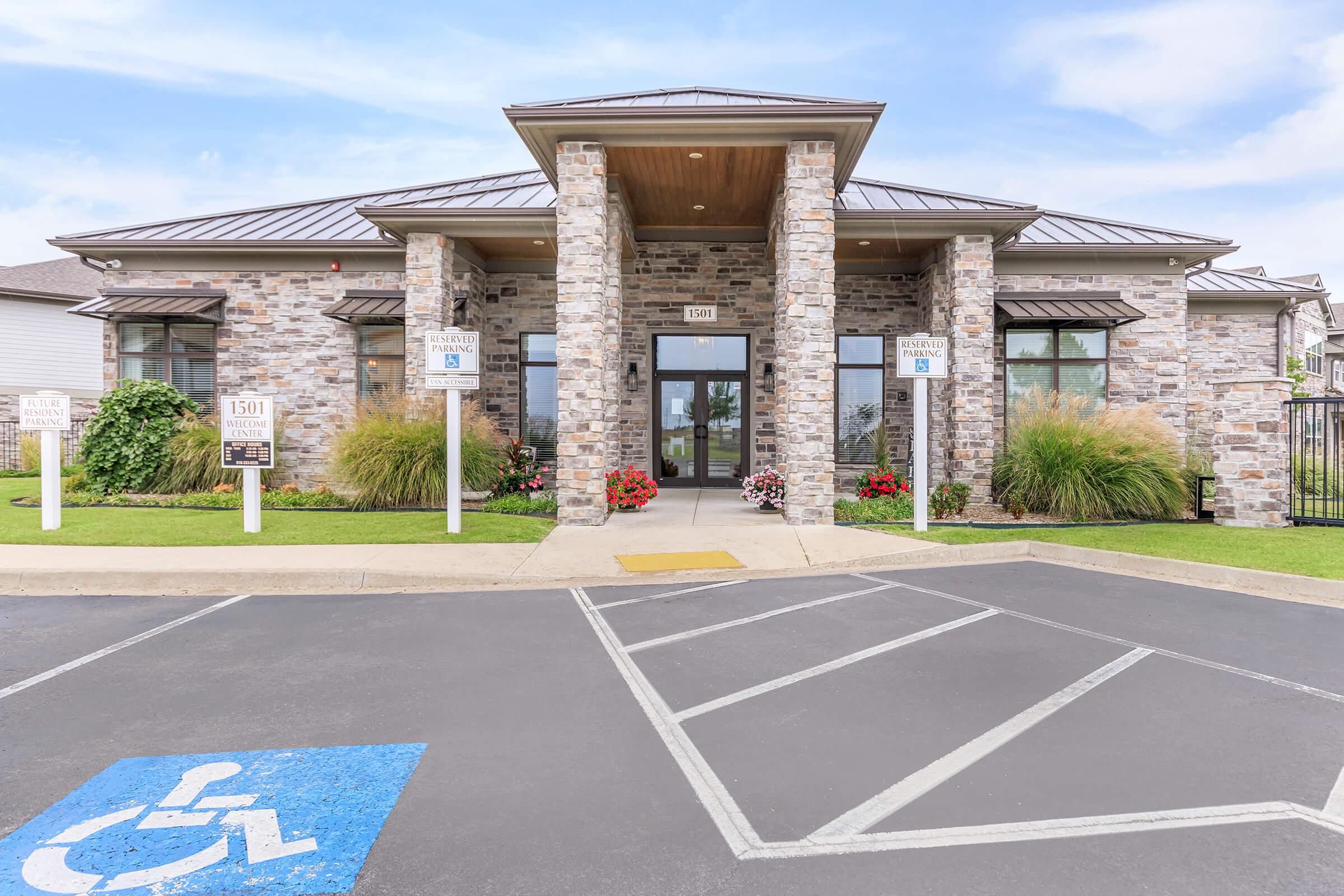A modern building with a stone façade and a peaked metal roof, featuring large glass doors at the entrance. The front is landscaped with ornamental grasses and flower beds. There is a designated handicapped parking space in the foreground, and additional signs nearby provide information about the location.