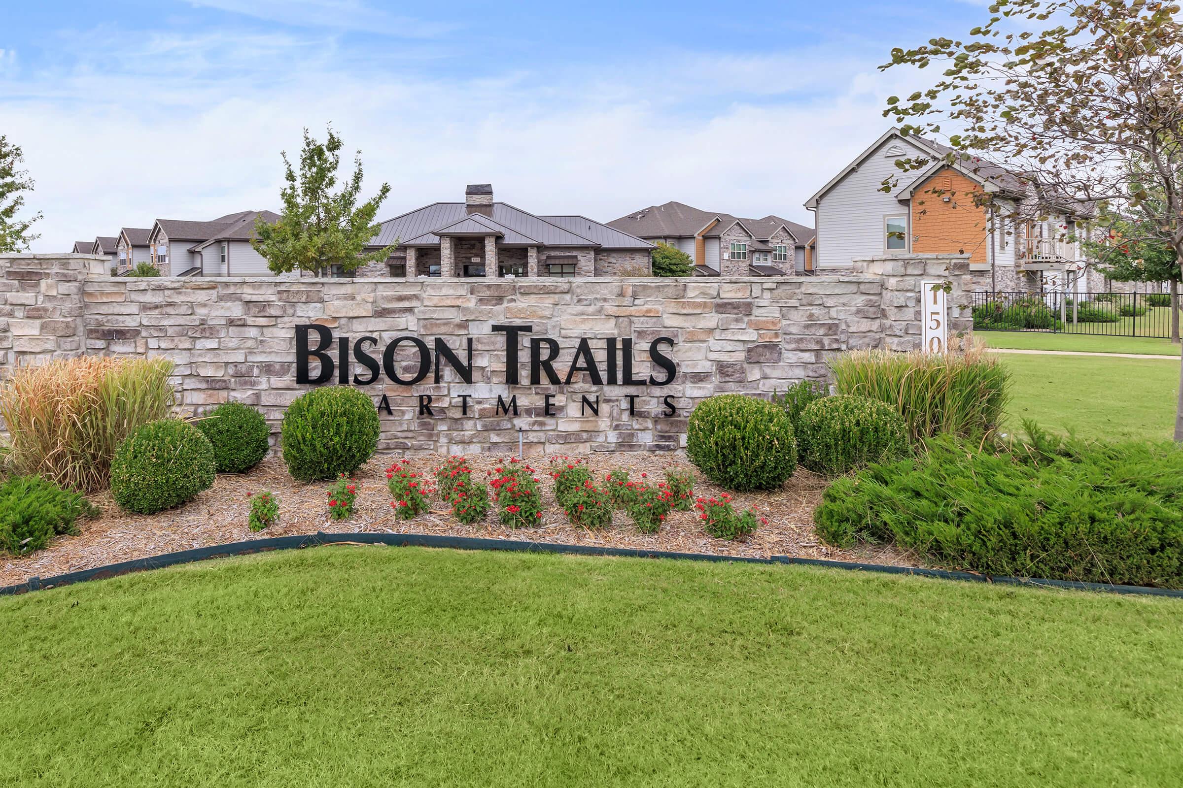 Sign for Bison Trails Apartments displaying the name prominently, set against a landscaped background with green grass, decorative bushes, and flower beds. In the background, modern residential buildings can be seen, and the sky is clear with a few clouds.