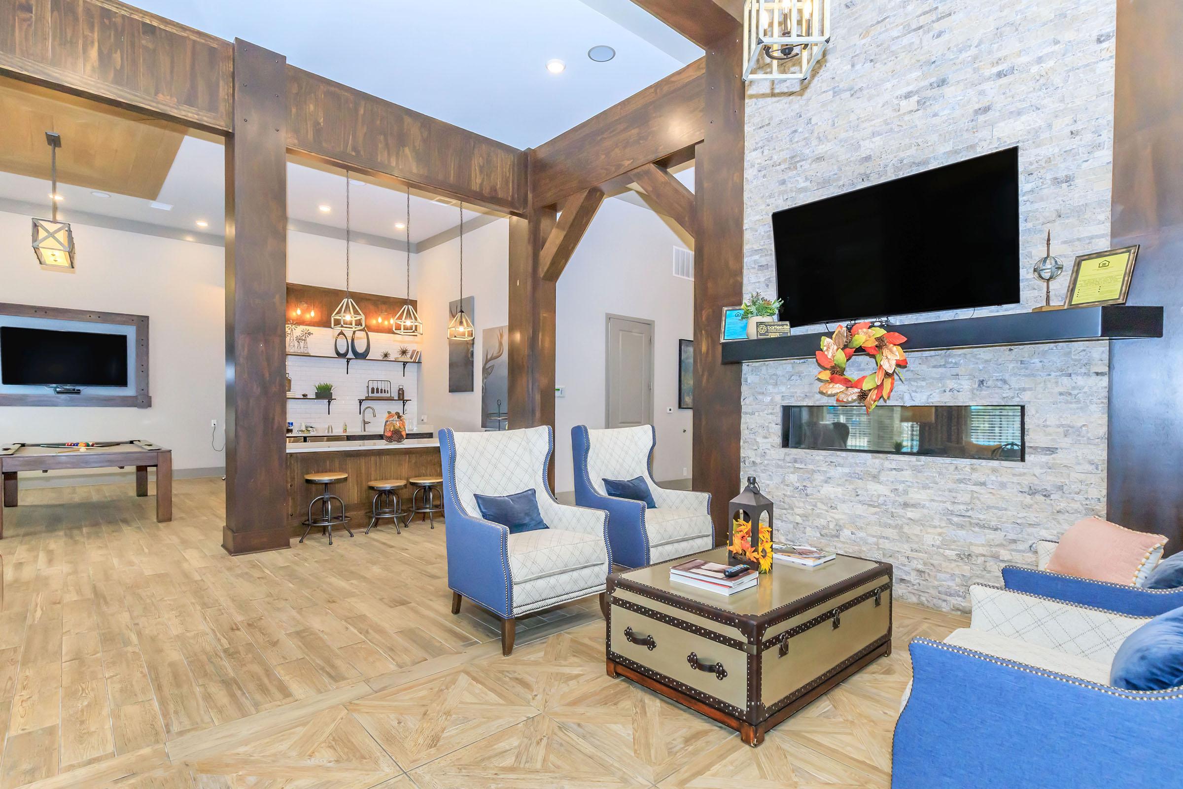 A cozy living area featuring two blue armchairs, a coffee table, and a large stone wall with a TV. The background shows a stylish kitchen with bar stools and decorative elements. Warm lighting and a touch of autumn decor complete the inviting atmosphere.