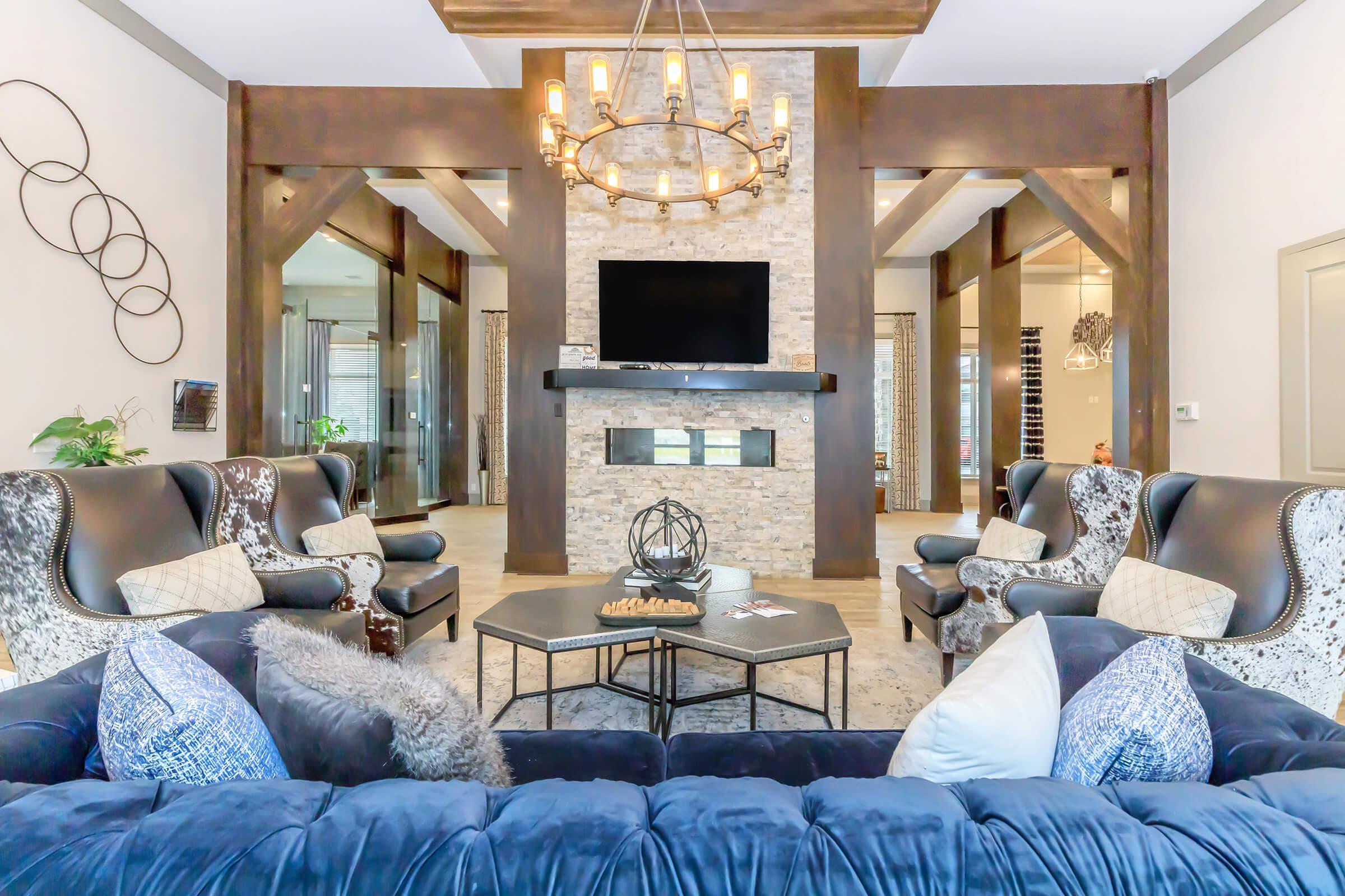 A spacious living room featuring two elegant armchairs and a plush blue couch with decorative pillows. A modern chandelier hangs from the ceiling, and a stone fireplace with a TV above it is the focal point. The room is warmly lit, with tall windows and contemporary decor adding to its inviting ambiance.