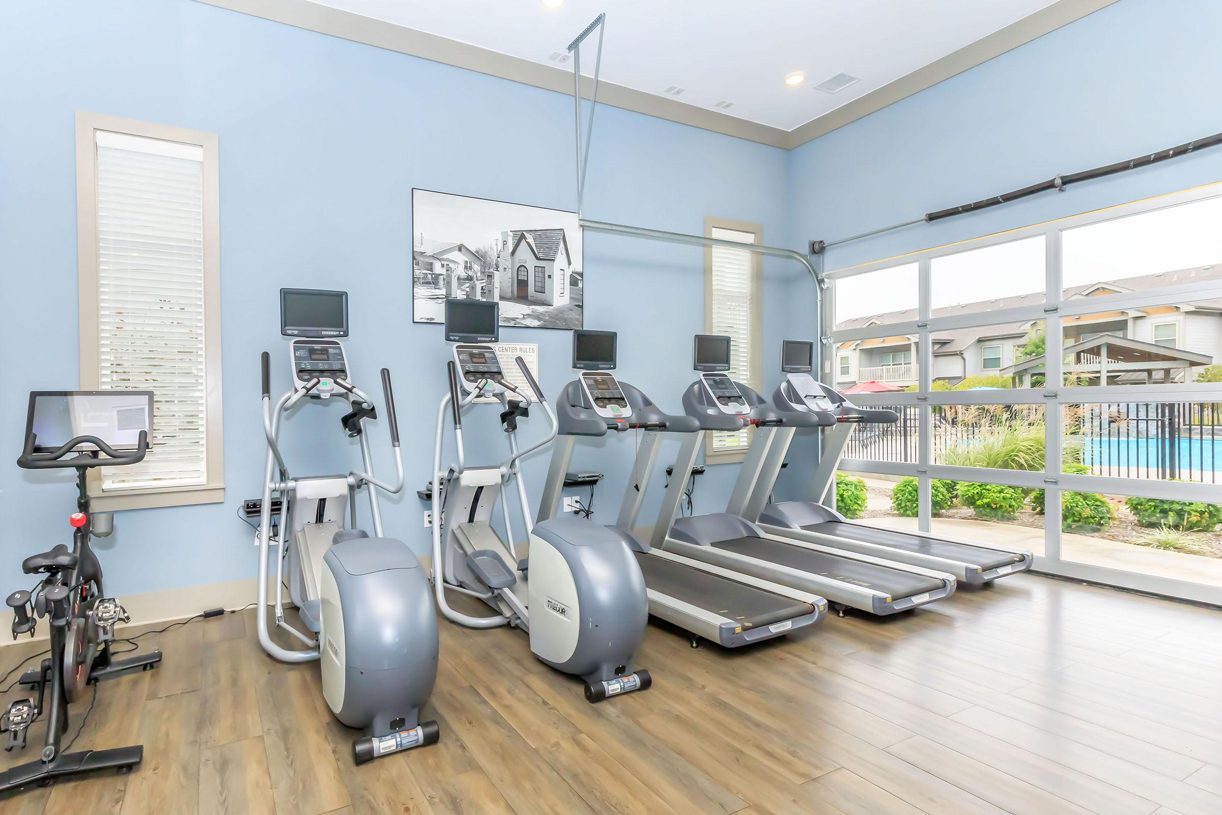 A modern gym interior featuring several exercise machines including treadmills, ellipticals, and a stationary bike, set against a light blue wall. Large windows allow natural light to fill the space, with a view of an outdoor pool area in the background.