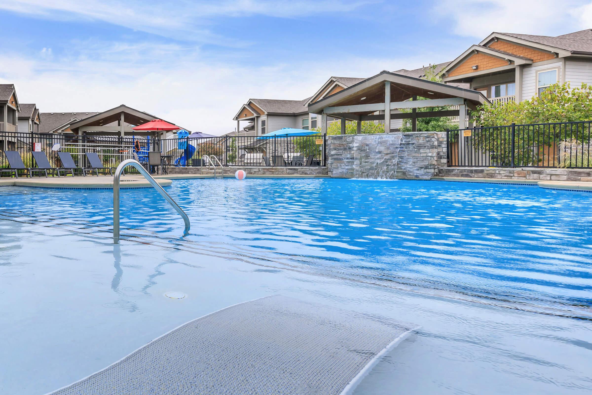 A clear blue swimming pool with a gentle incline and a handrail, surrounded by lounge chairs. In the background, there are colorful umbrellas and a gazebo, with modern residential buildings visible. The sky is bright and sunny, reflecting off the water's surface.