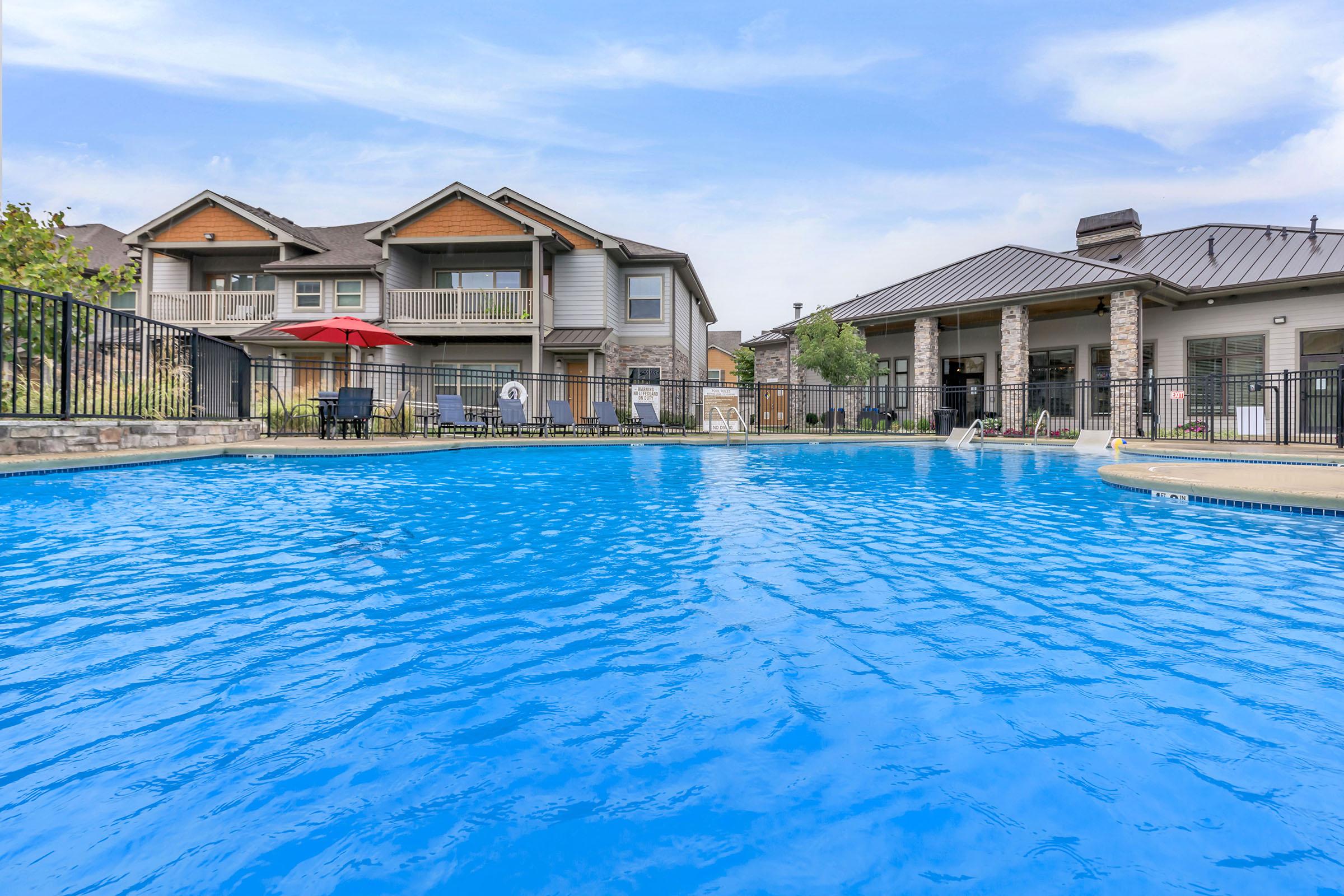 A clear blue swimming pool surrounded by a black fence, with sun loungers and a red umbrella. In the background, there are two multi-story buildings, one featuring a porch. The scene is bright and inviting, with a clear sky above.