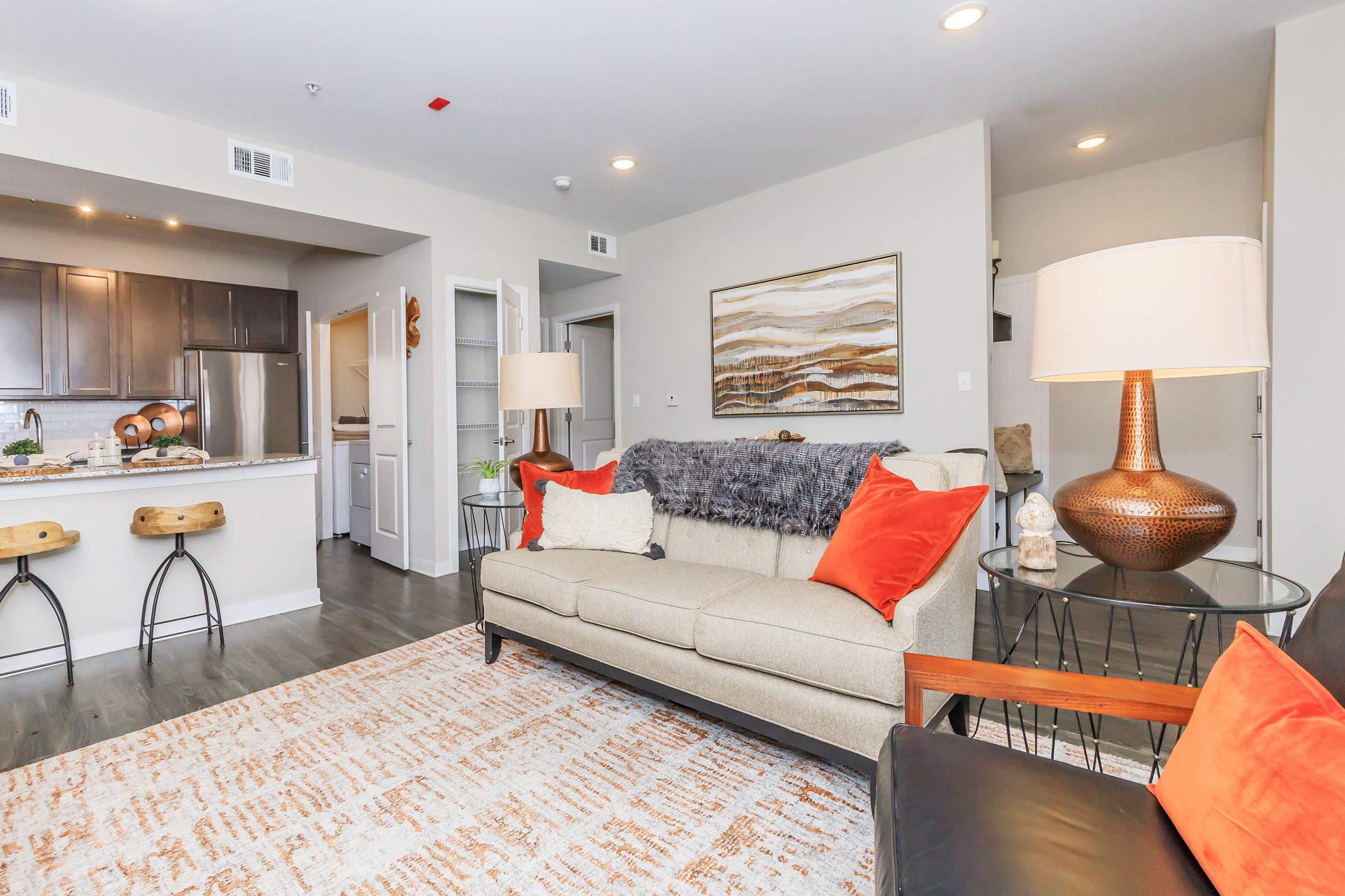 A cozy living room featuring a beige sofa with orange pillows, a black armchair, a stylish lamp, and a decorative rug. In the background, there is an open kitchen with dark cabinets and a dining area. Natural light fills the space, creating a warm and inviting atmosphere.