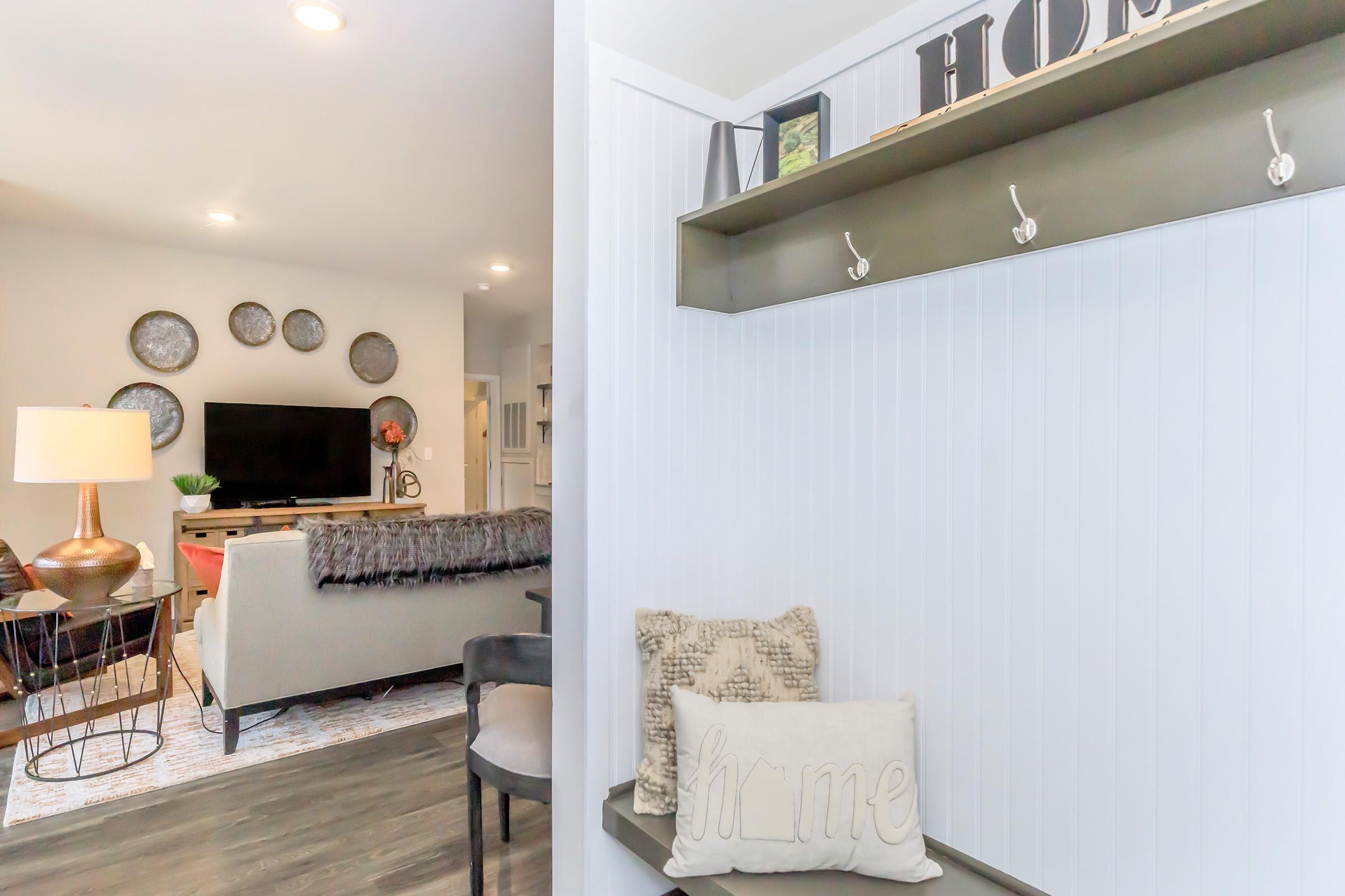 A cozy entryway leading to a modern living room. The entry features a wall-mounted coat rack, decorative pillows, and a warm lamp. The living area showcases a large TV, stylish furniture, and decorative wall plates, with soft lighting creating an inviting atmosphere.
