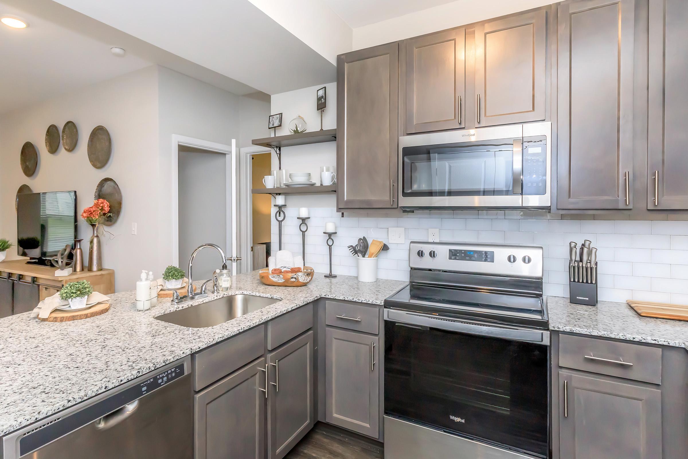 A modern kitchen featuring dark wooden cabinets, a stainless steel oven and microwave, a granite countertop, and a double sink. Decorative items, including plants and dishware, are arranged on the counter. The space is well-lit and has a clean, contemporary aesthetic.