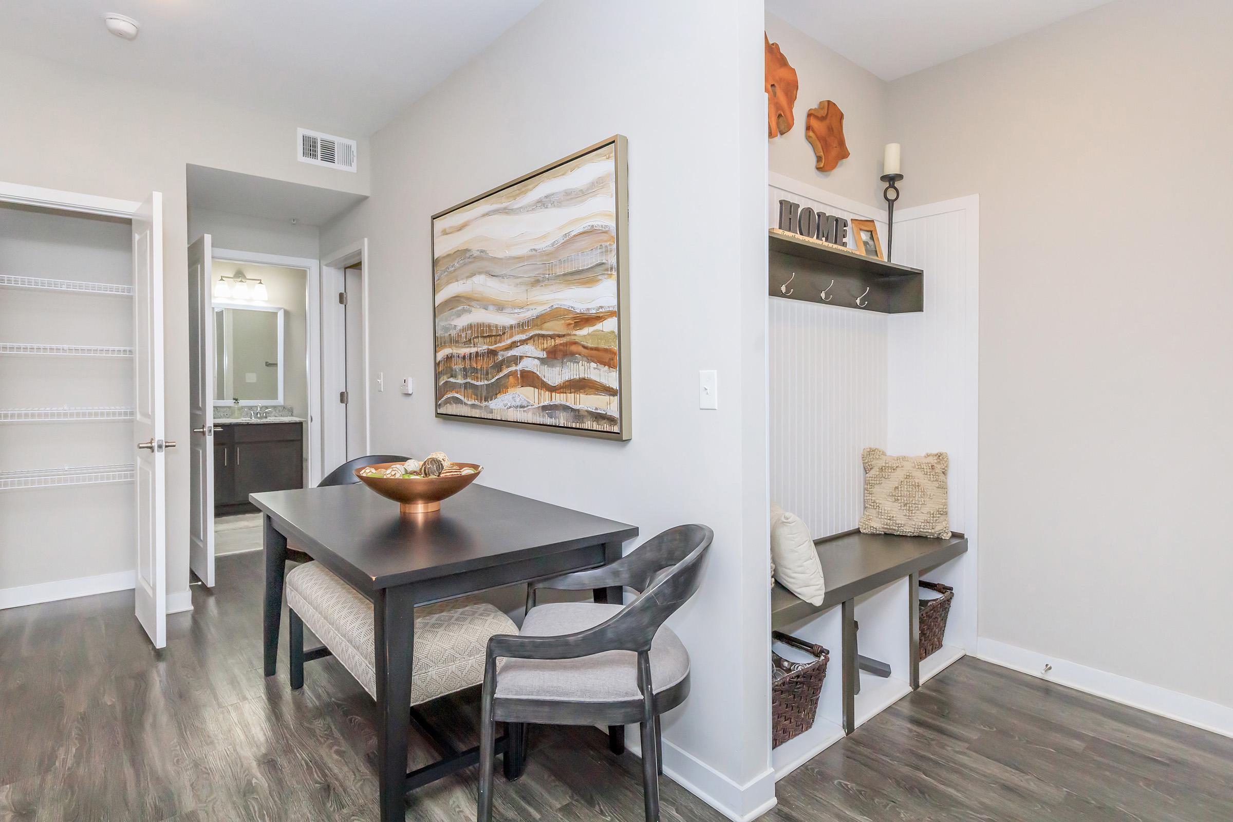 A modern dining area featuring a sleek black table with gray chairs, near a decorative wall art piece. To the side, there's a small entryway with a bench and baskets, and a glimpse of a doorway leading to a bathroom. The walls are painted in neutral tones, creating a bright and inviting atmosphere.