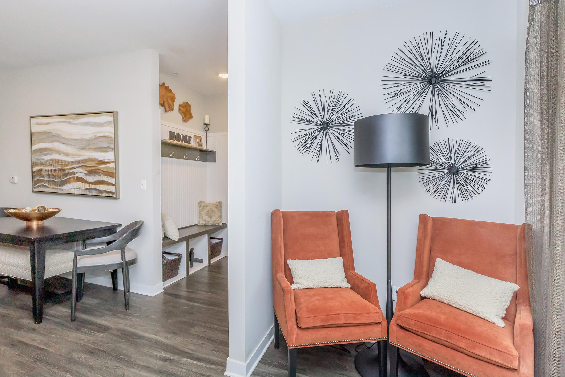 Two orange armchairs with decorative pillows are positioned in a modern living space. A dark floor lamp stands beside them. In the background, a dining area features a wooden table with gray chairs. The wall displays three decorative starburst designs, and a contrasting artwork hangs nearby.