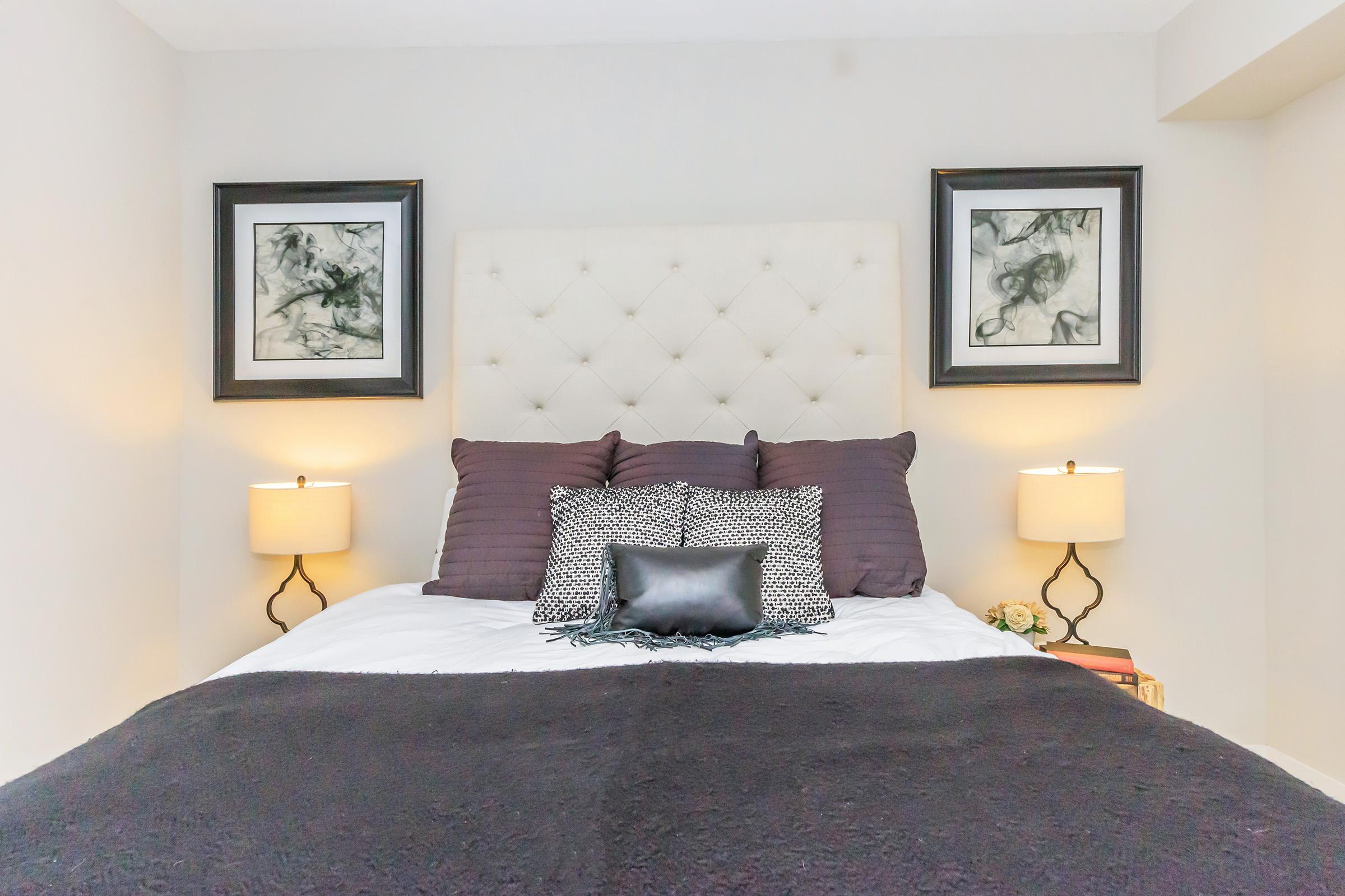 A well-decorated bedroom featuring a light-colored tufted headboard with dark purple pillows, flanked by framed abstract artwork. Two bedside lamps provide soft lighting on either side, and a dark throw blanket covers the bed. A small decorative pillow adds a touch of elegance, creating a cozy atmosphere.