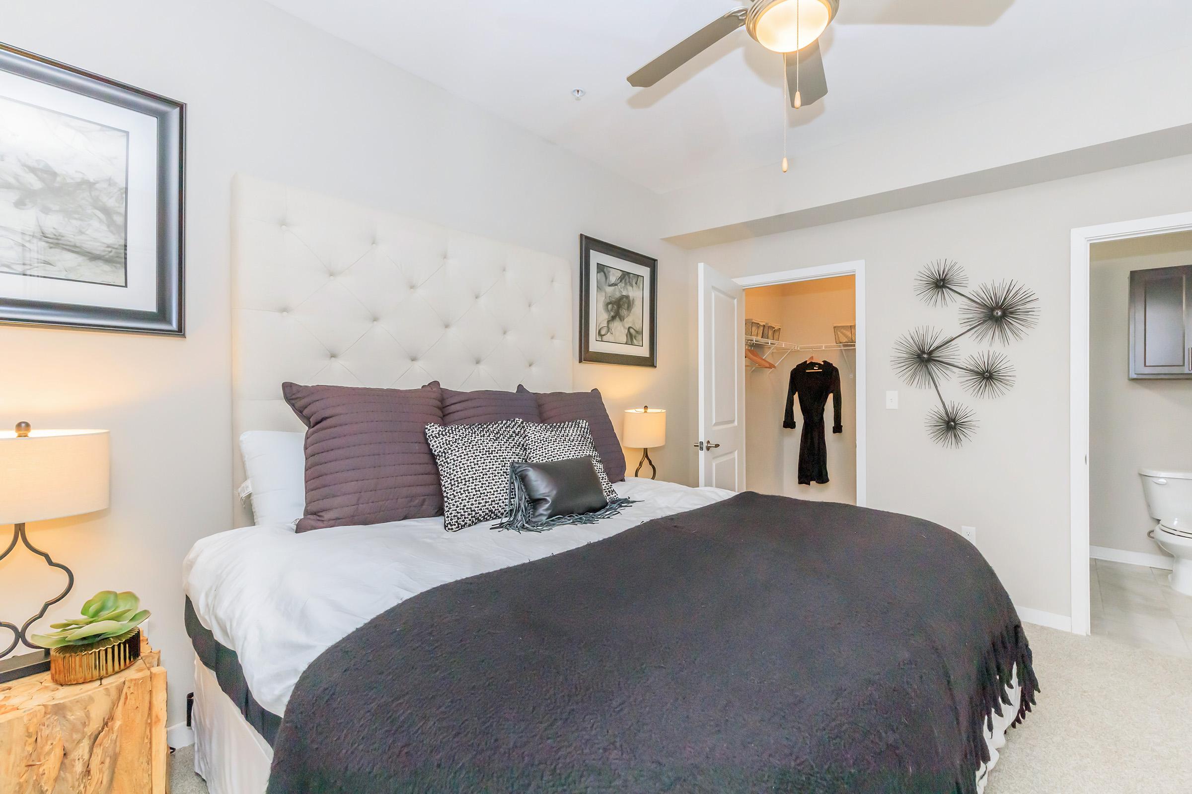 A modern bedroom featuring a plush bed with gray and black bedding, decorative pillows, and a stylish headboard. The room has tasteful artwork on the walls, bedside lamps on wooden nightstands, and a walk-in closet visible in the background. A ceiling fan adds to the contemporary decor.