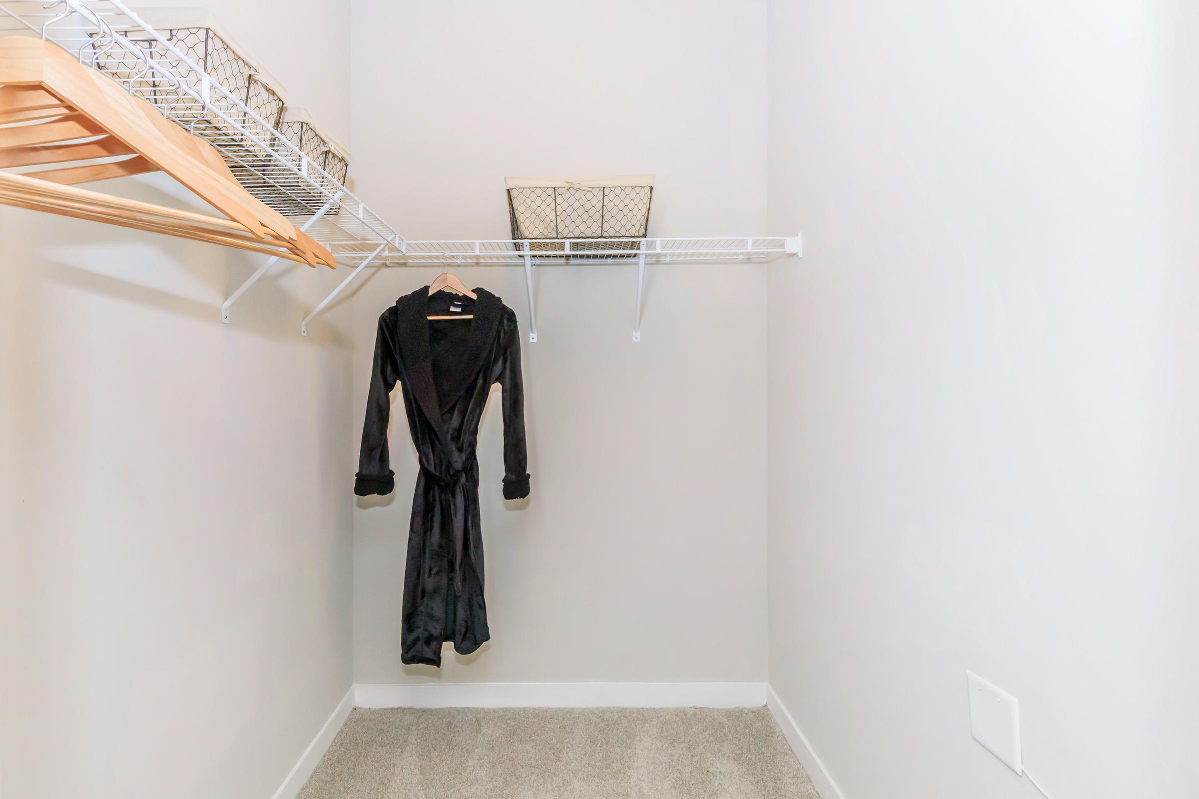 A minimalistic closet featuring a hanging black robe on a wire shelf, with empty wooden hangers and a couple of woven baskets. The walls are painted a light color, and the floor is carpeted in a neutral tone.