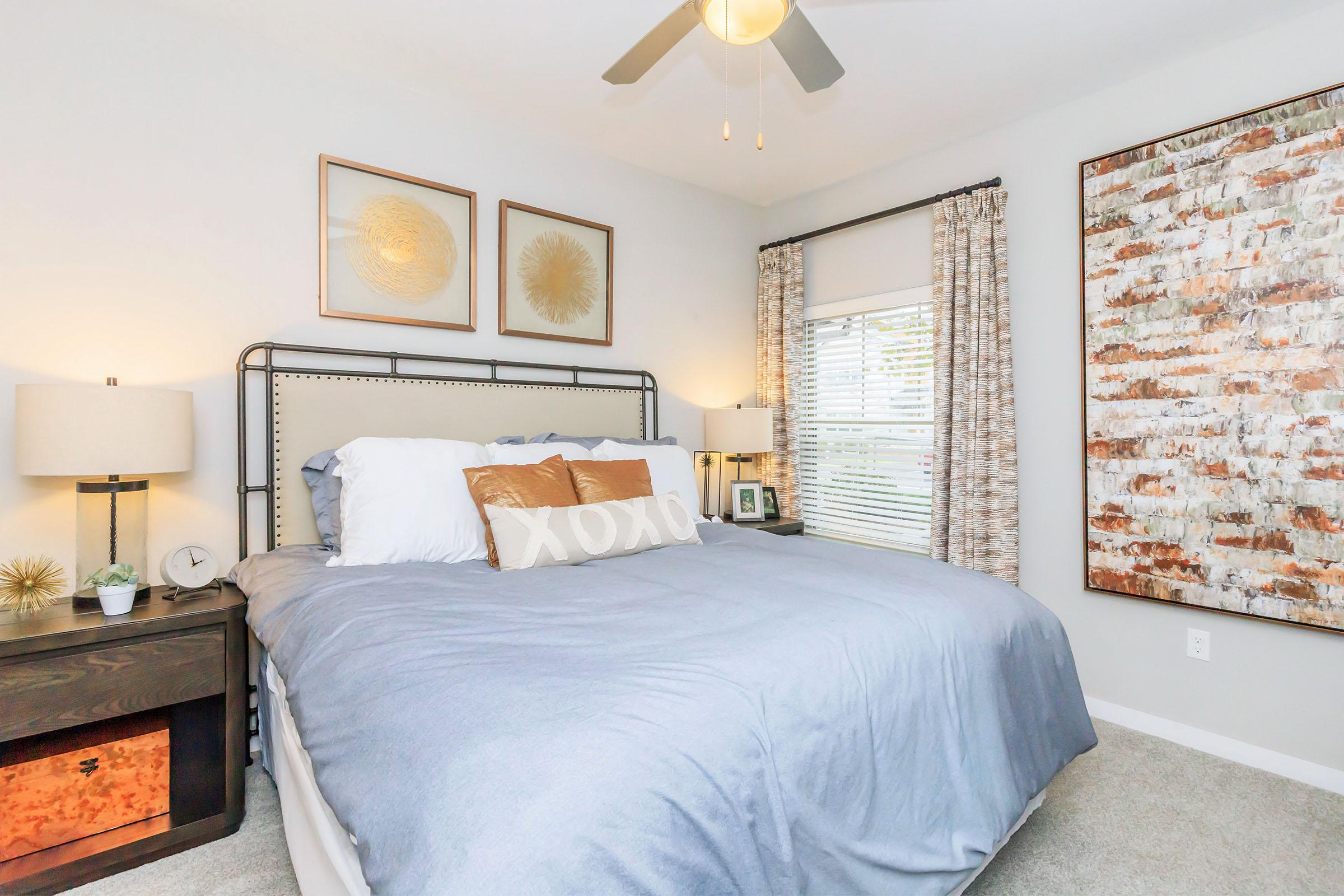 A cozy bedroom featuring a large bed with blue bedding and decorative pillows. There are two bedside lamps and a nightstand with a clock. The walls are adorned with framed artwork and a textured panel. Natural light filters through window treatments, creating a warm atmosphere.