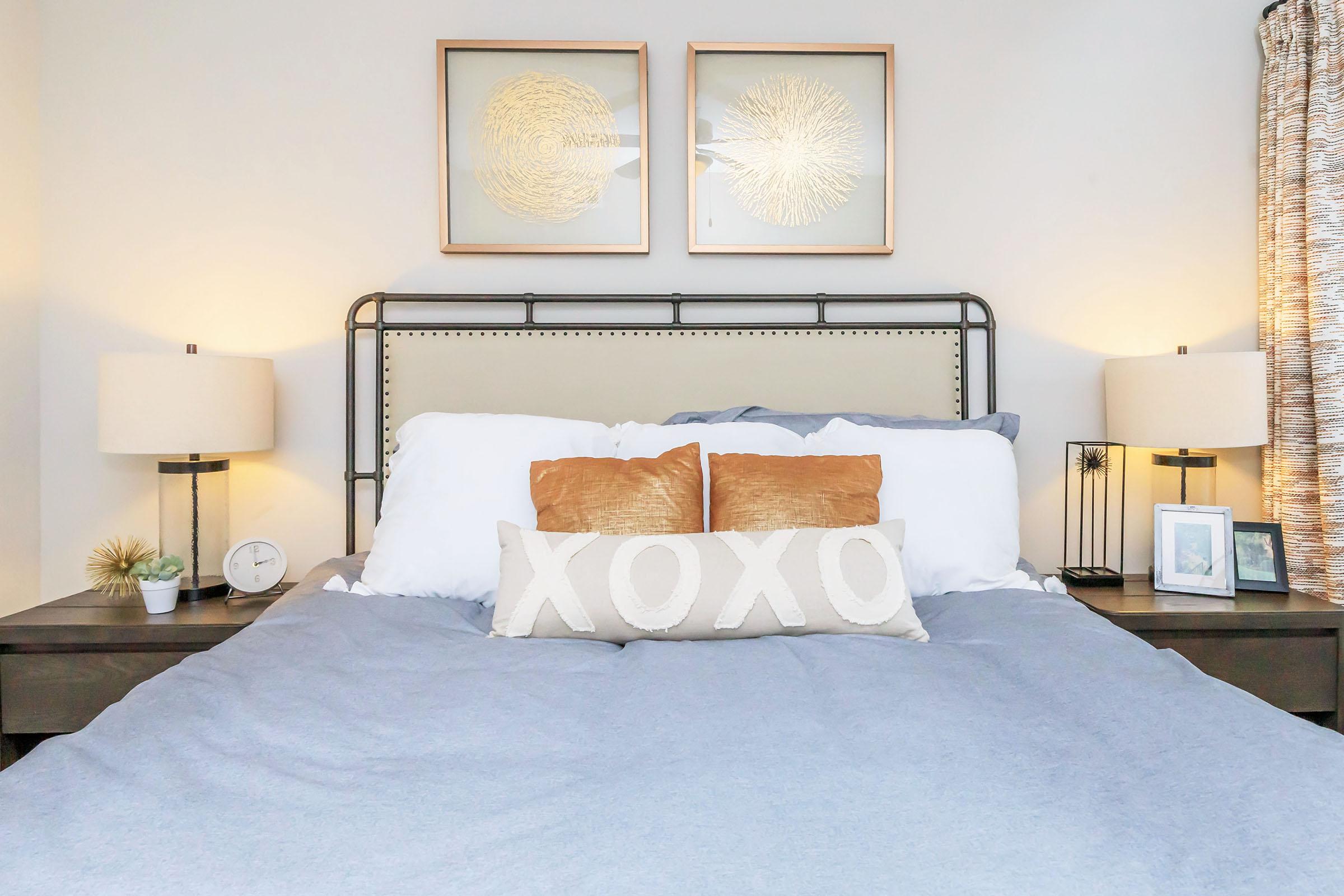 A cozy bedroom with a metal bed frame and a large, soft bedding in blue. Two decorative pillows, one with the word "XOXO" in white, sit on the bed. There are two bedside lamps illuminating the space, a clock and a small plant on one nightstand, and framed art above the bed. A window with patterned curtains is visible.