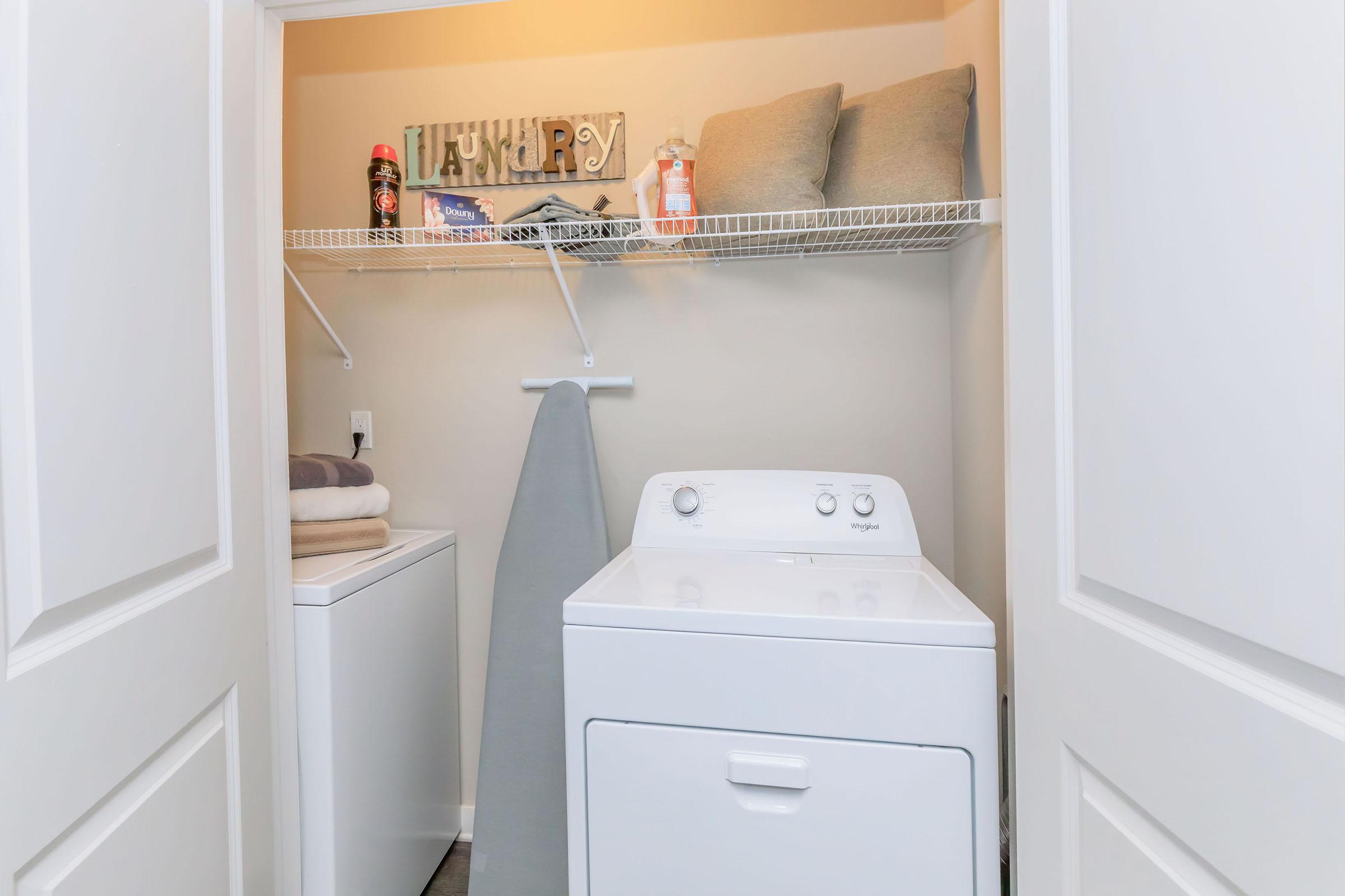A tidy laundry room featuring a white washing machine and dryer side by side. Above, shelves hold decorative items, bottles, and two gray pillows. An ironing board is propped against the wall, and stacked towels are neatly placed on the dryer. The walls are painted a neutral tone, creating a clean and organized space.