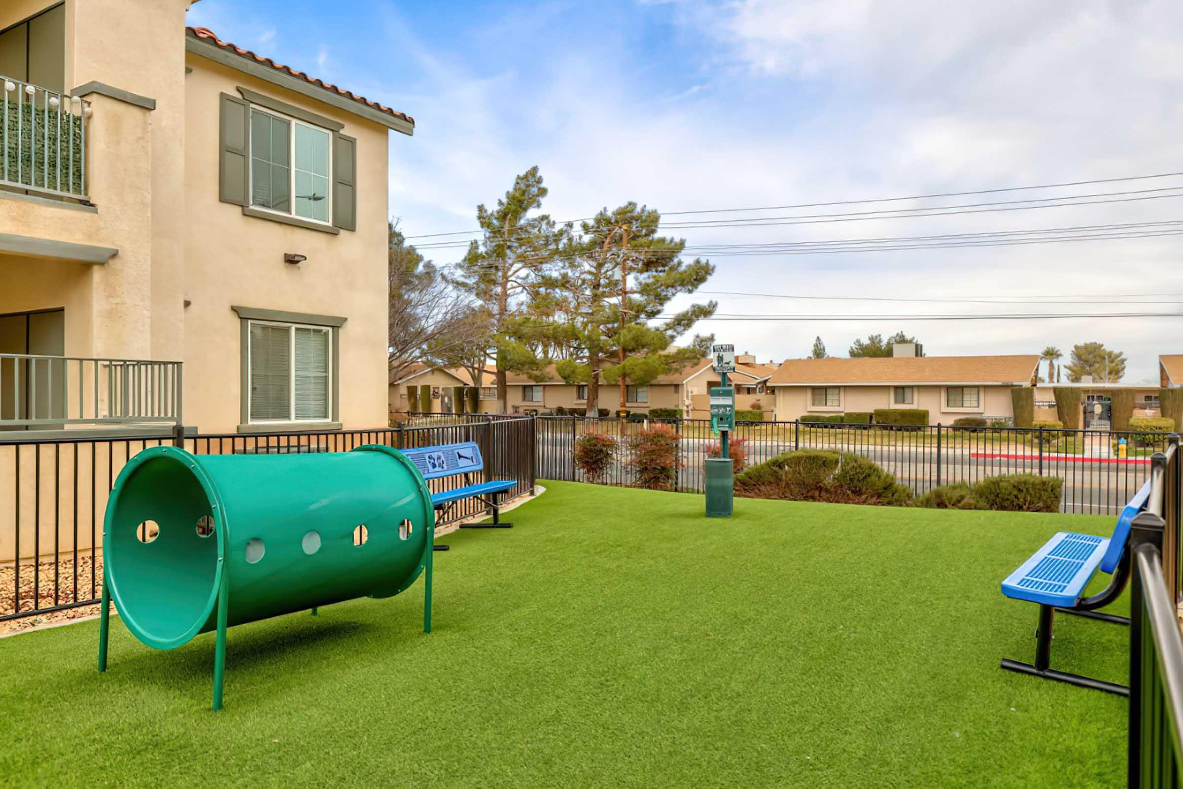 A small outdoor dog park area featuring a green tunnel, two blue benches, and artificial grass. The park is enclosed by a black fence, with a backdrop of apartment buildings and trees under a partly cloudy sky.