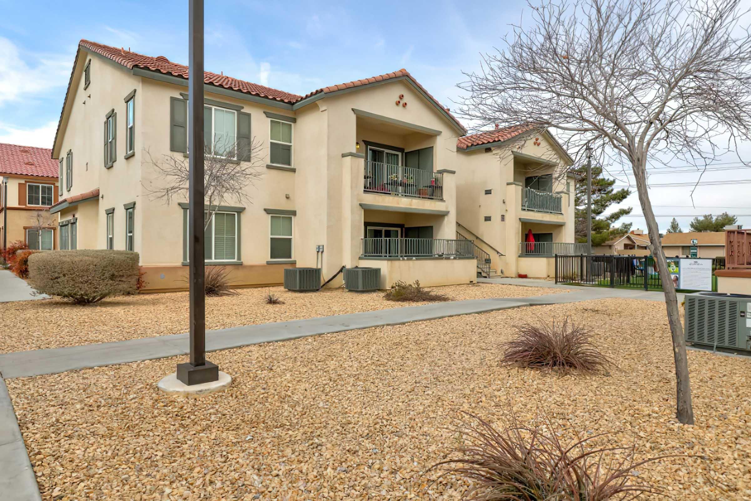 A view of a residential building with a light-colored exterior, featuring two stories and multiple balconies. The landscaping includes decorative gravel and sparse vegetation. There are air conditioning units visible, and pathways leading to the entrance. A clear sky can be seen in the background.