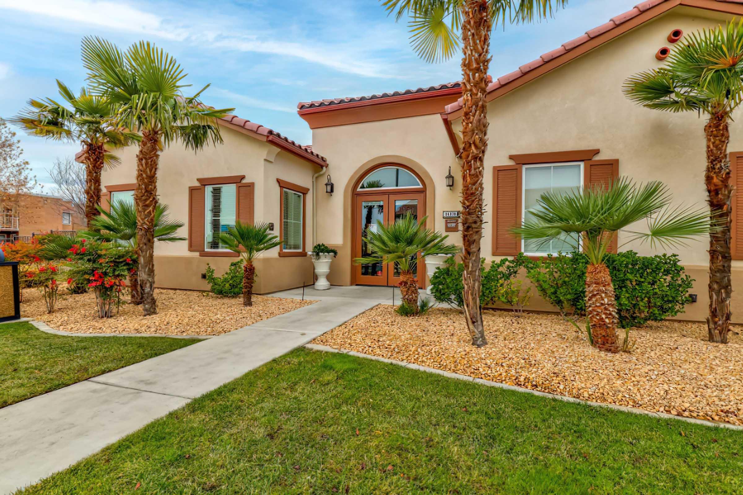 A beautiful single-story home with a tiled roof, surrounded by palm trees and well-maintained landscaping. The pathway leads to a welcoming entrance with large windows and decorative shutters, set against a clear blue sky.