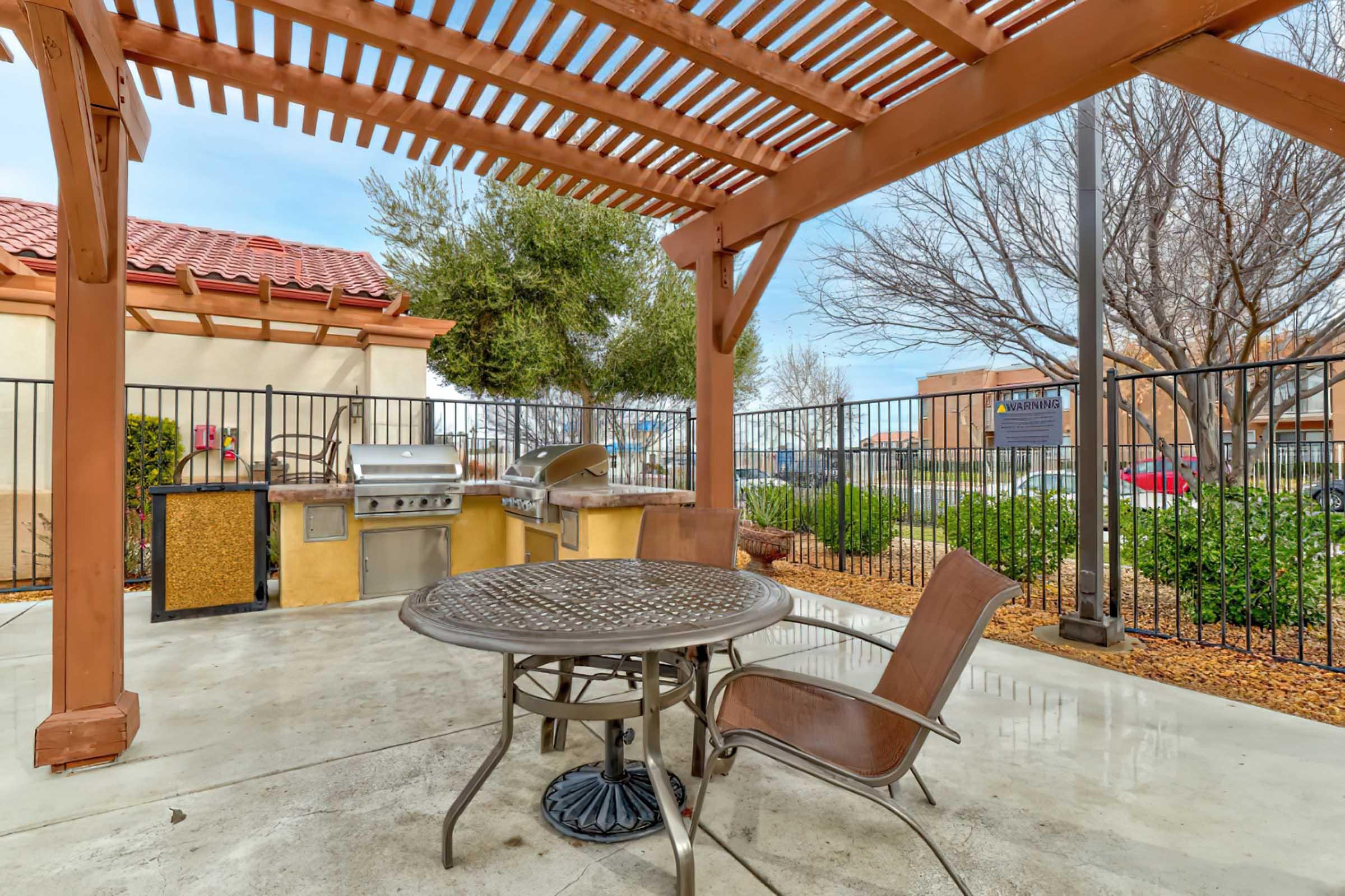 Outdoor patio area featuring a shaded pergola, a round table with chairs, and a built-in barbecue grill. The space is surrounded by low shrubs and decorative gravel, with a fence in the background. The scene is set against a clear sky and nearby buildings.