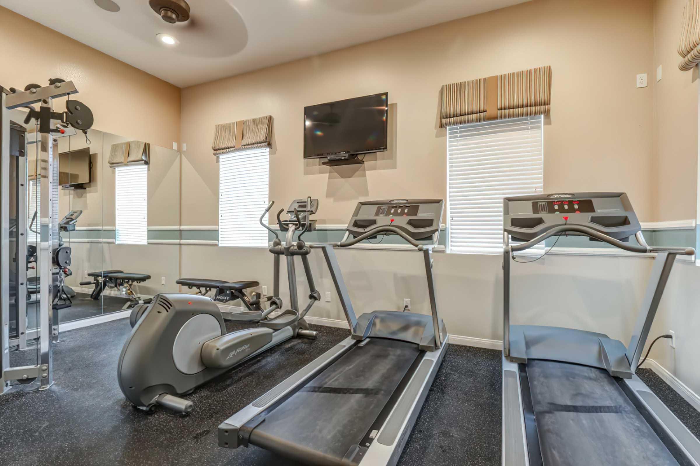 A fitness room featuring two treadmills, a stationary bike, and a multi-gym weight machine. The space has a mirror, windows with blinds, and a wall-mounted television, all set against a neutral-colored wall with a black rubber floor.