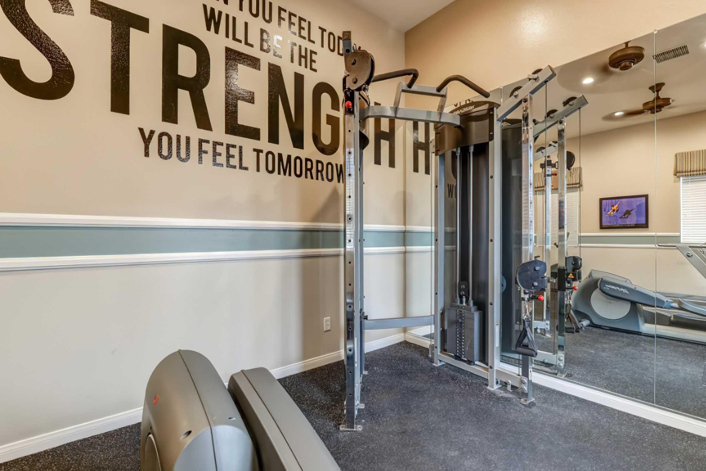 A home gym with a cable machine and exercise equipment. The walls feature motivational text about strength, with a mirror on one side reflecting the gym setup. The space is well-lit and organized, emphasizing fitness and wellbeing.